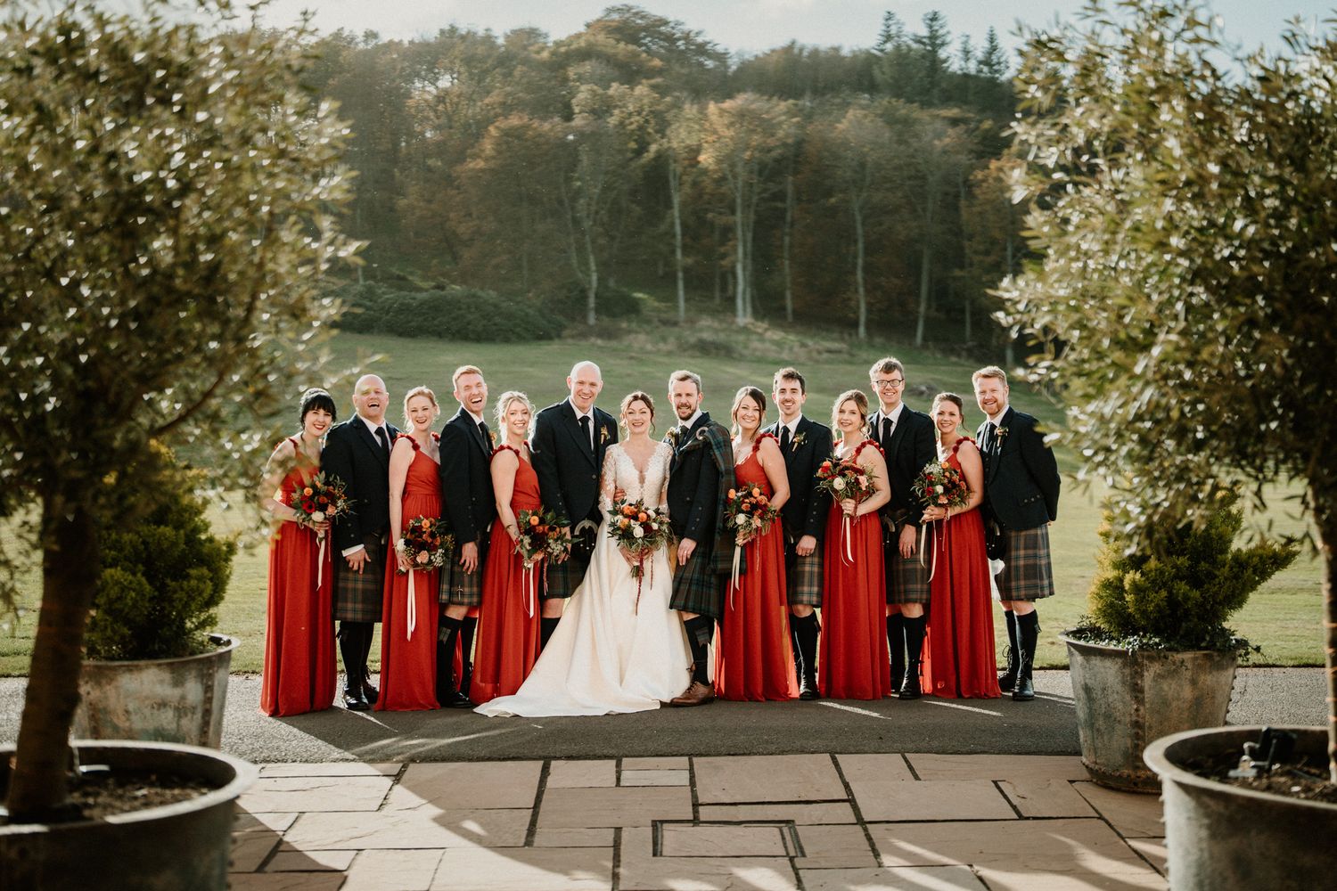 Panoramic wedding party photo with bridesmaids in red dresses against scenic landscape.