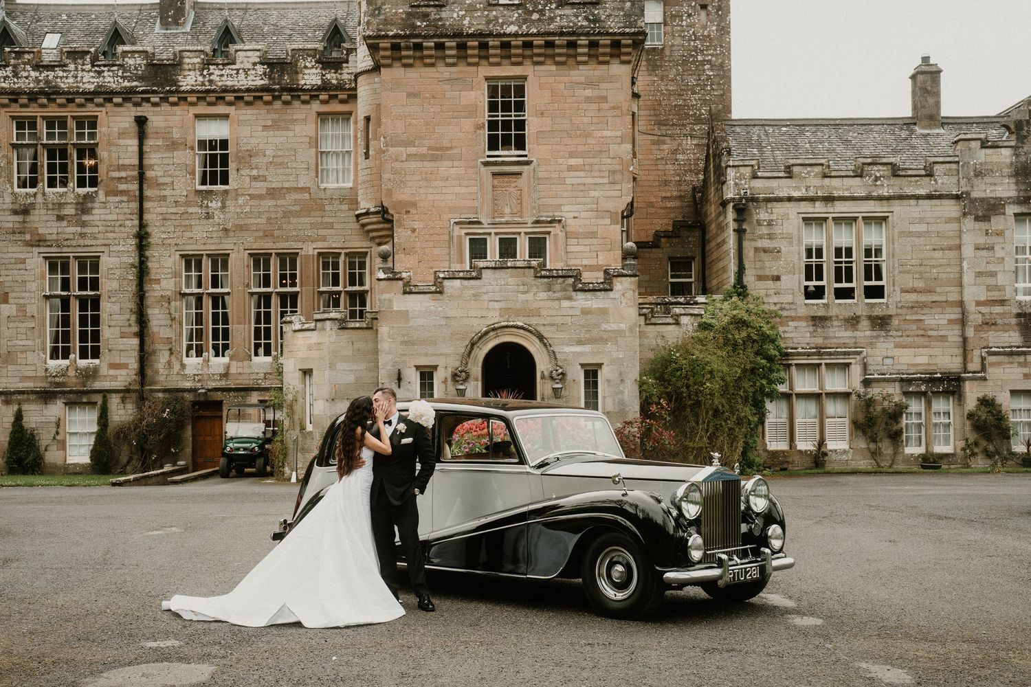 A wedding couple poses with a classic vintage car outside a grand stone castle estate.