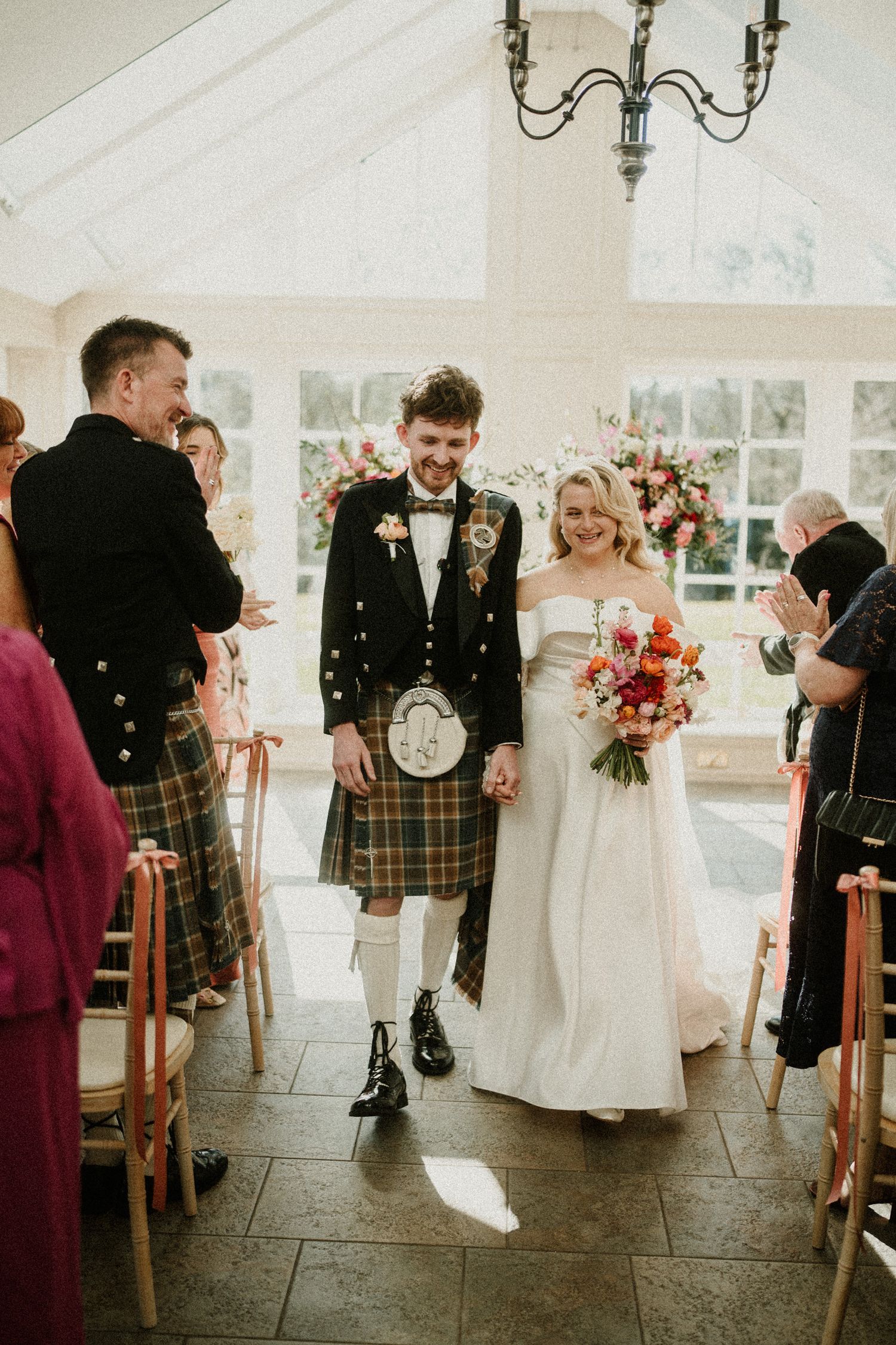 Wedding couple exits ceremony venue, groom wearing traditional Scottish attire and bride in white dress.