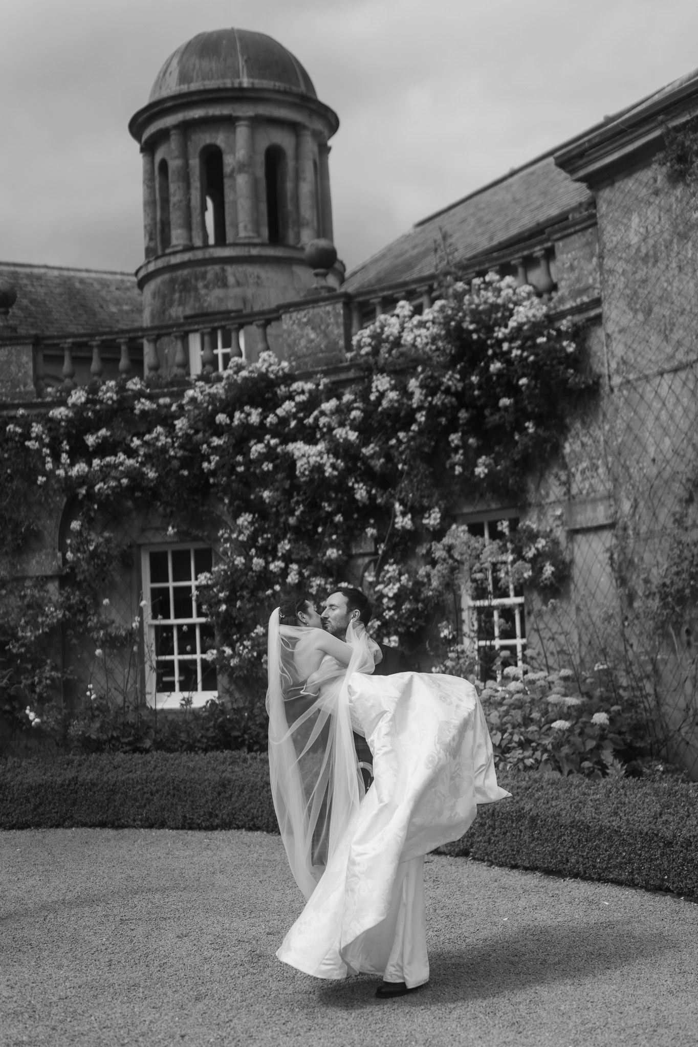 Black and white photo of a bride in flowing gown and veil outside a historic stone manor with climbing roses.