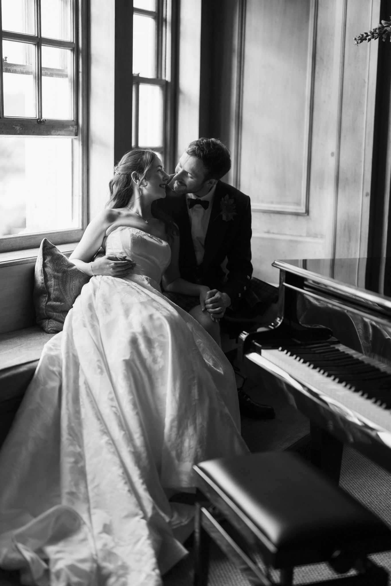 Black and white photo of a bride and groom sharing a romantic moment beside a grand piano near a window.