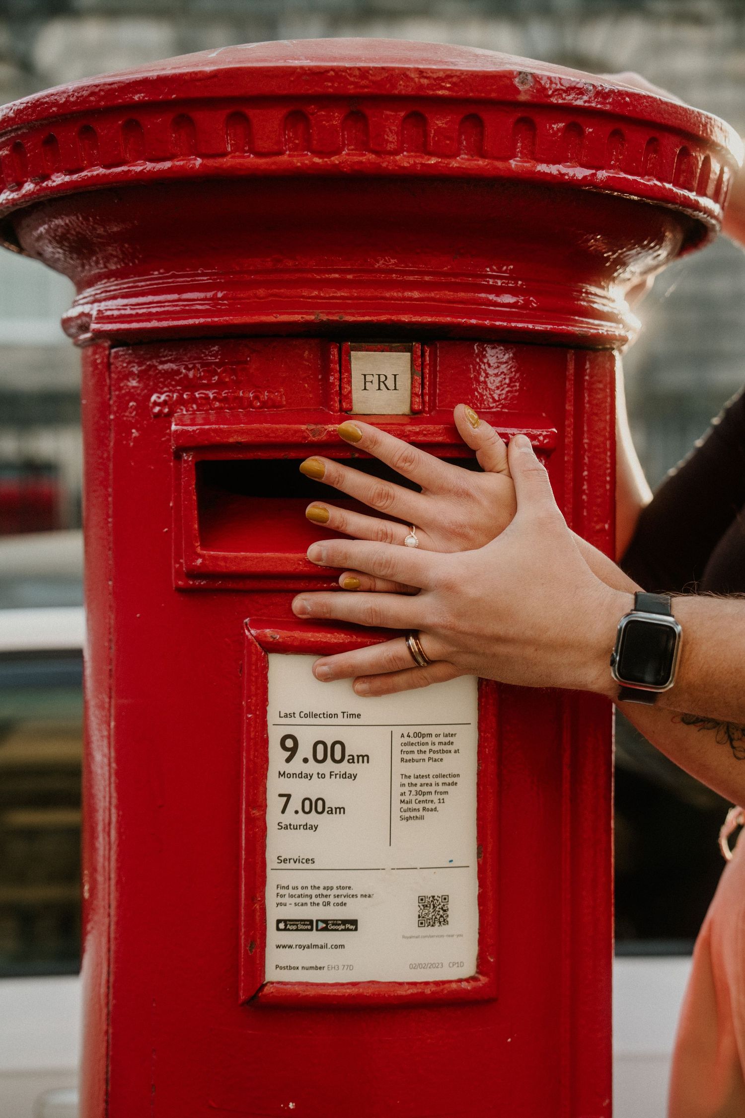 Hands posting a letter into a iconic red British Royal Mail pillar box with collection times visible.