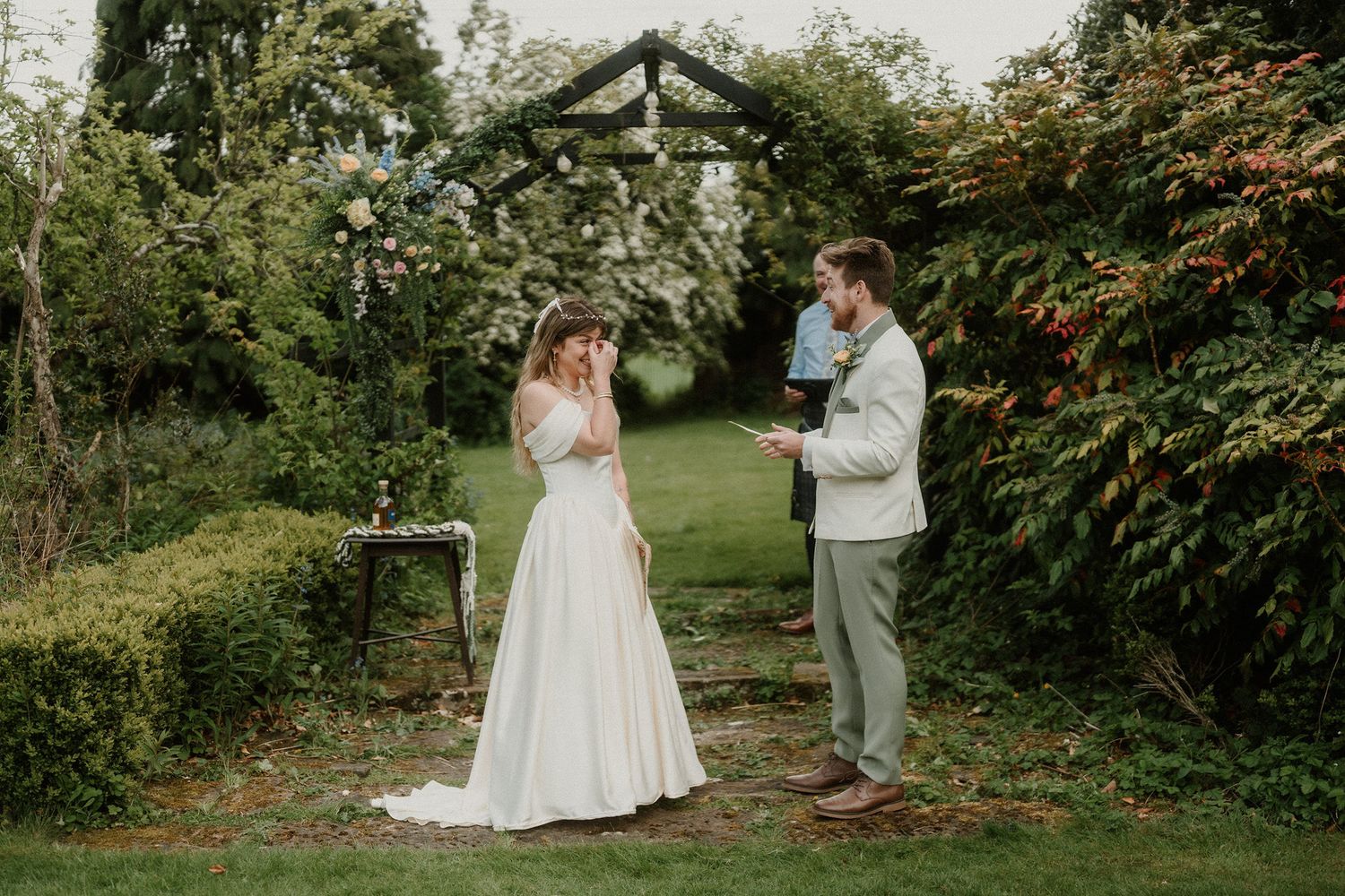 Couple exchanging vows under floral arch in lush garden wedding ceremony, bride in white gown, groom in grey suit.