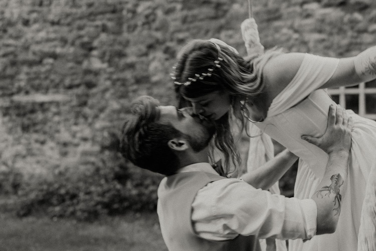 Black and white photo of a groom lifting his bride for a romantic kiss outdoors near a stone wall.