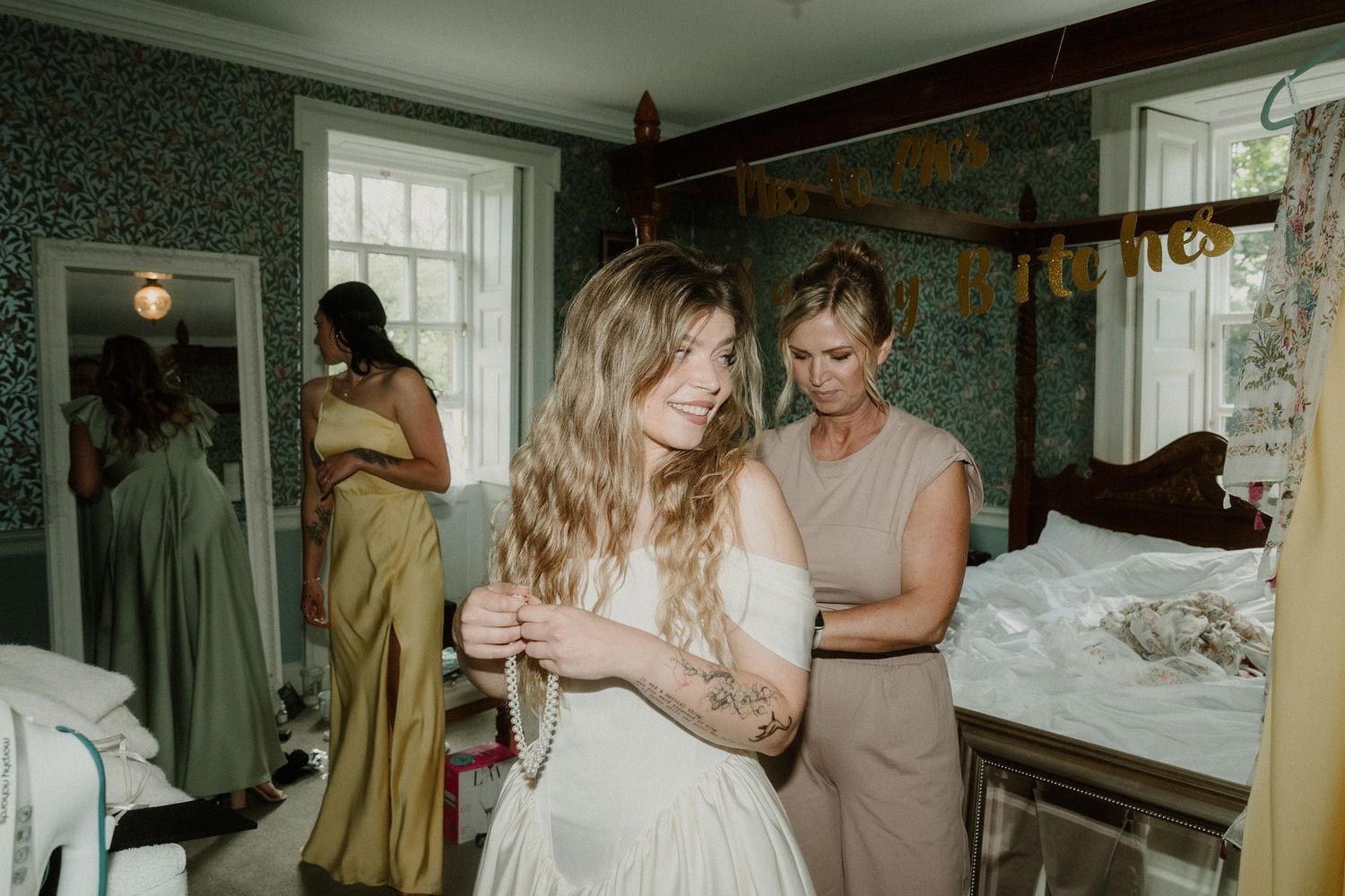 Bride getting dressed with help from bridesmaid in elegant green wallpapered bedroom on wedding day.