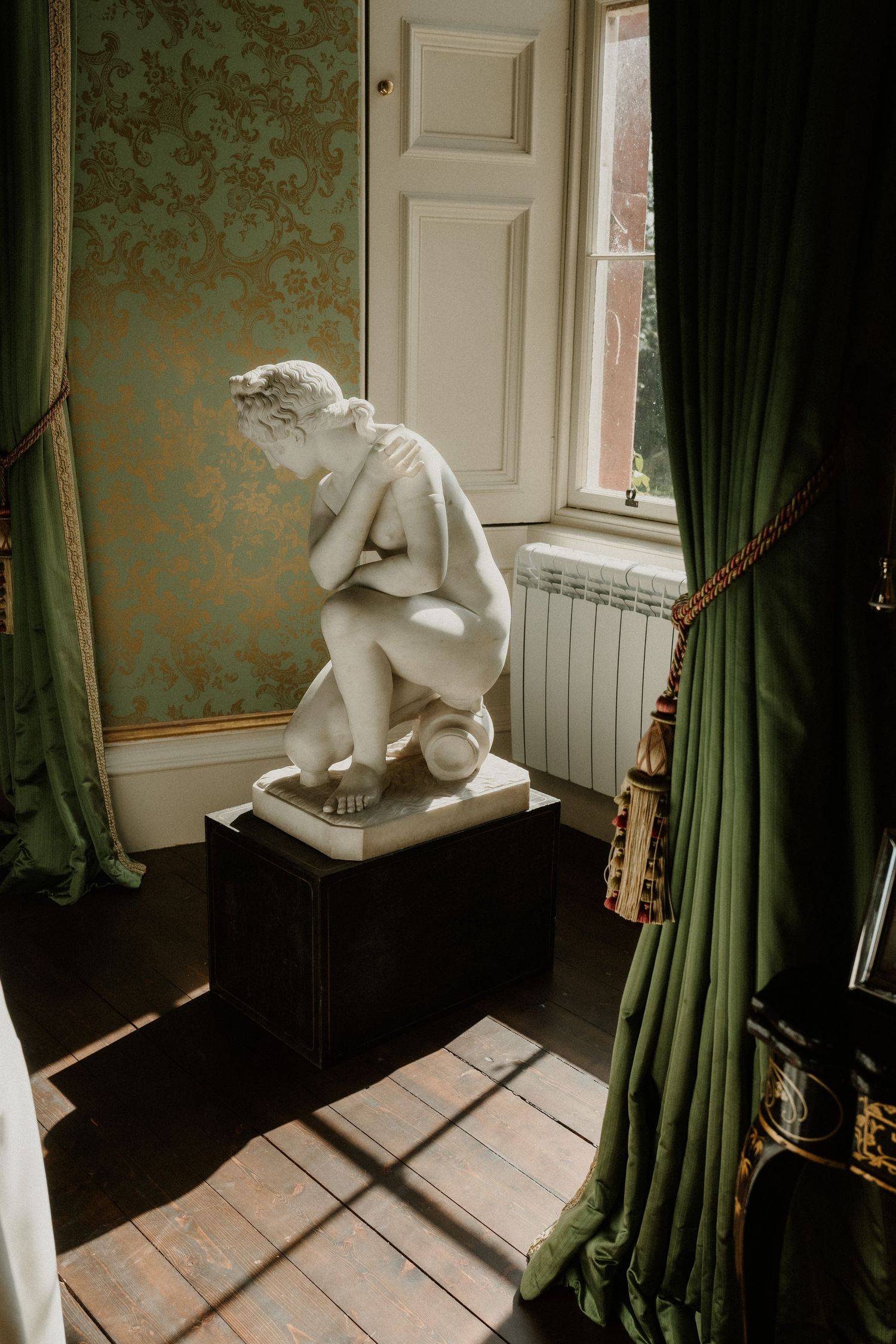 White marble crouching nude statue on black pedestal in elegant room with green damask walls and velvet curtains.