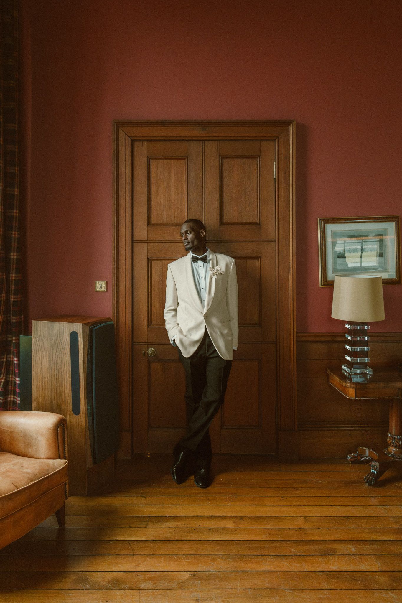 A person in a white jacket stands against a wooden-paneled wall in a room with warm reddish lighting and hardwood floors.