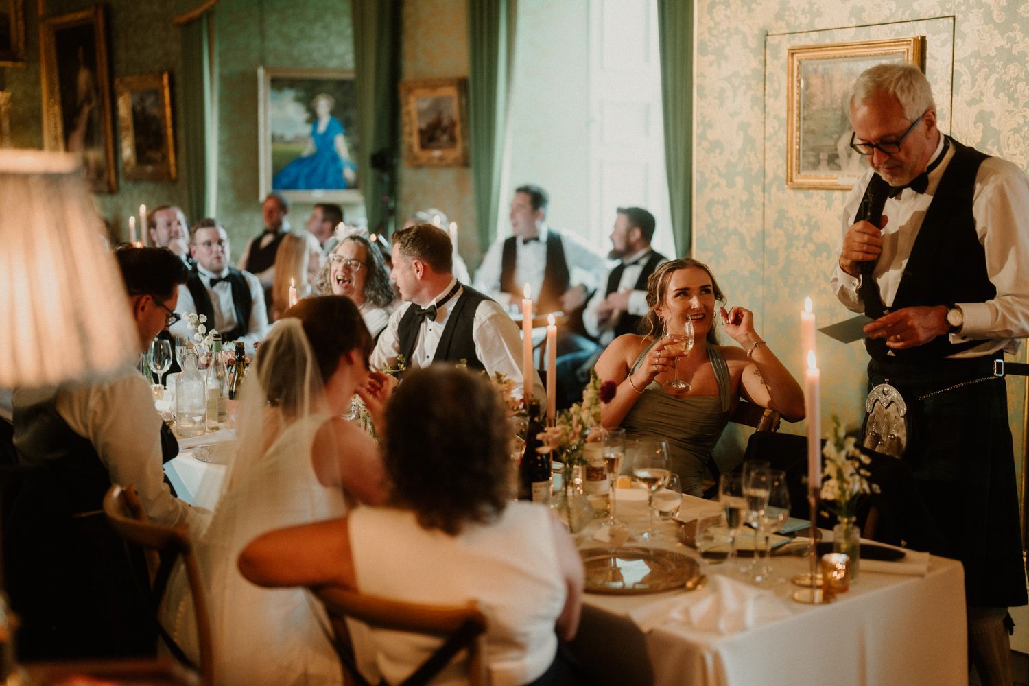 Man in a waistcoat and bow tie speaks into a microphone at a candlelit wedding reception while seated guests laugh and listen.