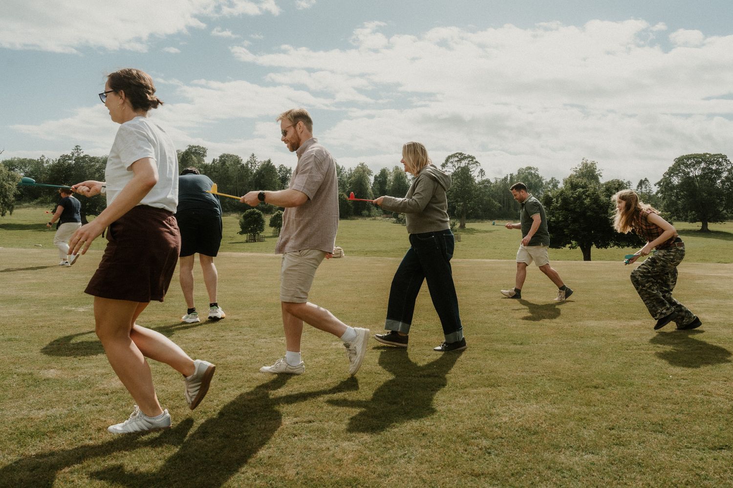 A group of people playing tug of war on a sunny grass field with trees and a partly cloudy sky in the background.