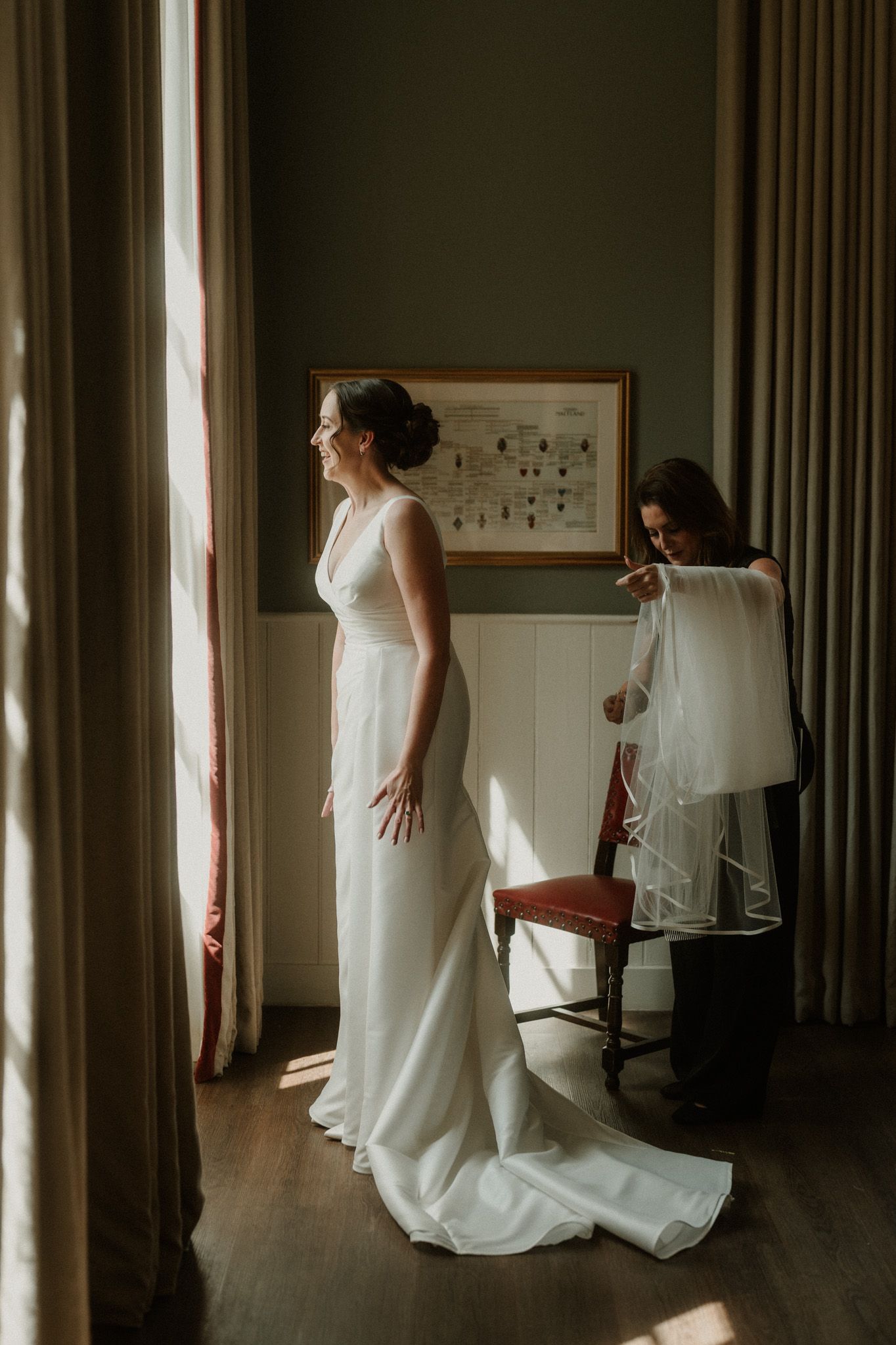 Bride in an elegant white gown with a long train stands by a sunlit window while a woman prepares her veil in a classic room.