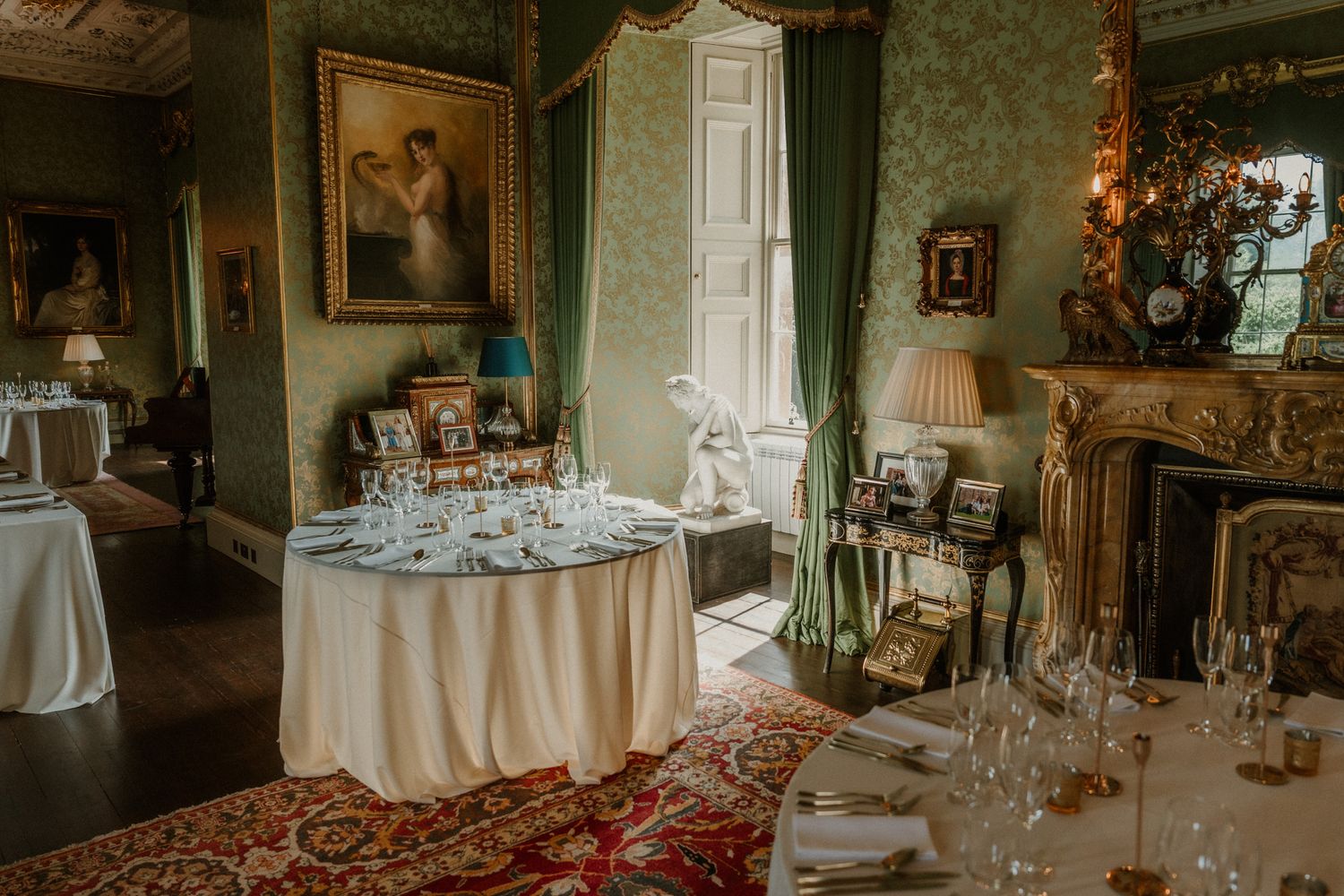 Elegant historic dining room with round tables set with glassware, green damask walls, ornate fireplace, framed portrait and Persian rug.