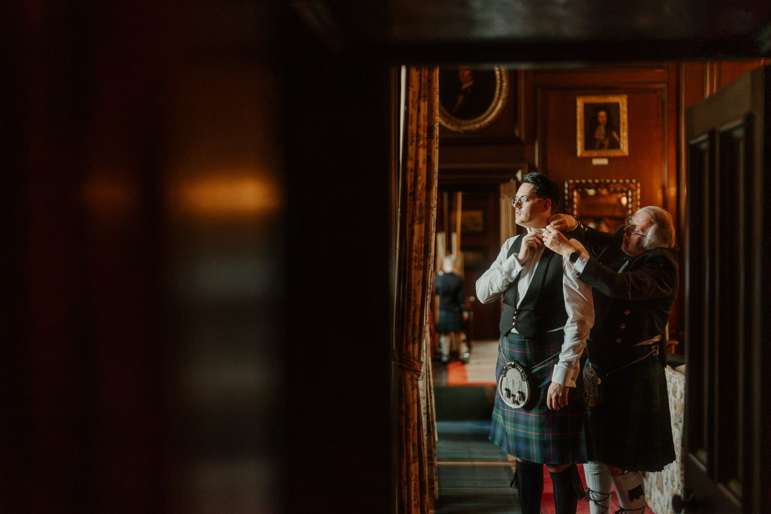 Groom in a green tartan kilt and waistcoat has his attire adjusted by a man in a warmly lit, wood-paneled room with framed portraits.