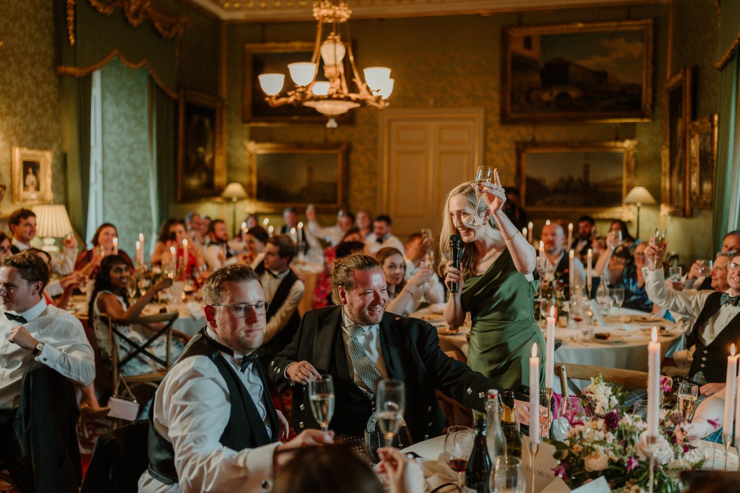 Wedding guests laugh and cheer at candlelit tables during a toast in an elegant ballroom with a chandelier and a woman in a green dress.