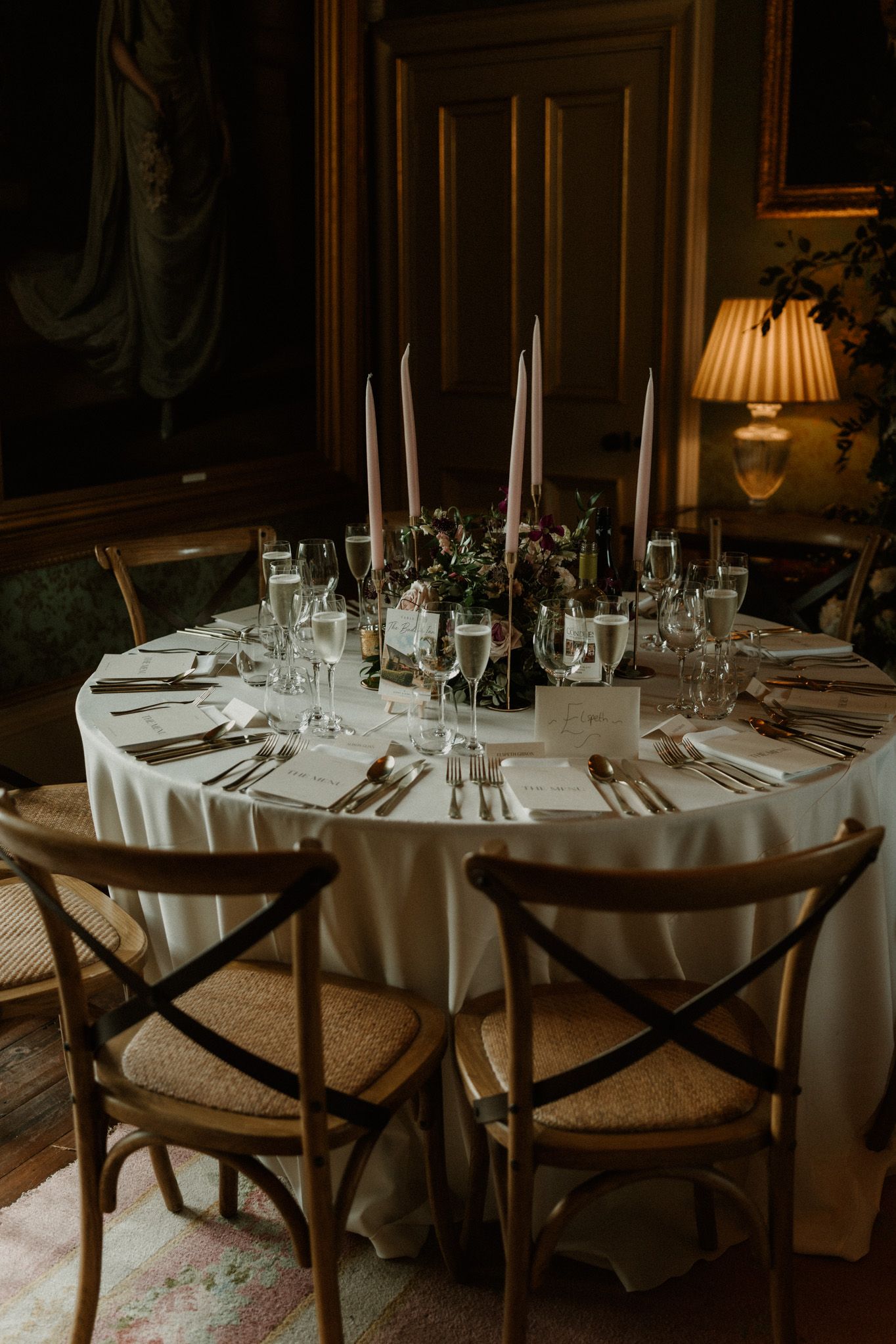 Elegant round wedding reception table set with white linens, tall taper candles, wine glasses, and floral centerpiece in a candlelit room.