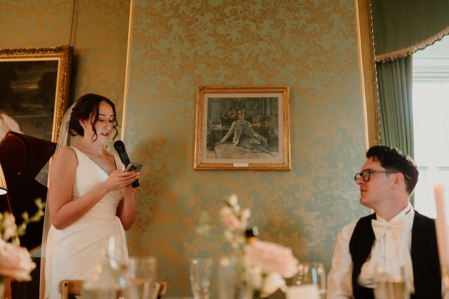 Woman in a white dress reads a wedding speech into a microphone in an ornate green room while a bearded man in glasses listens at the table.
