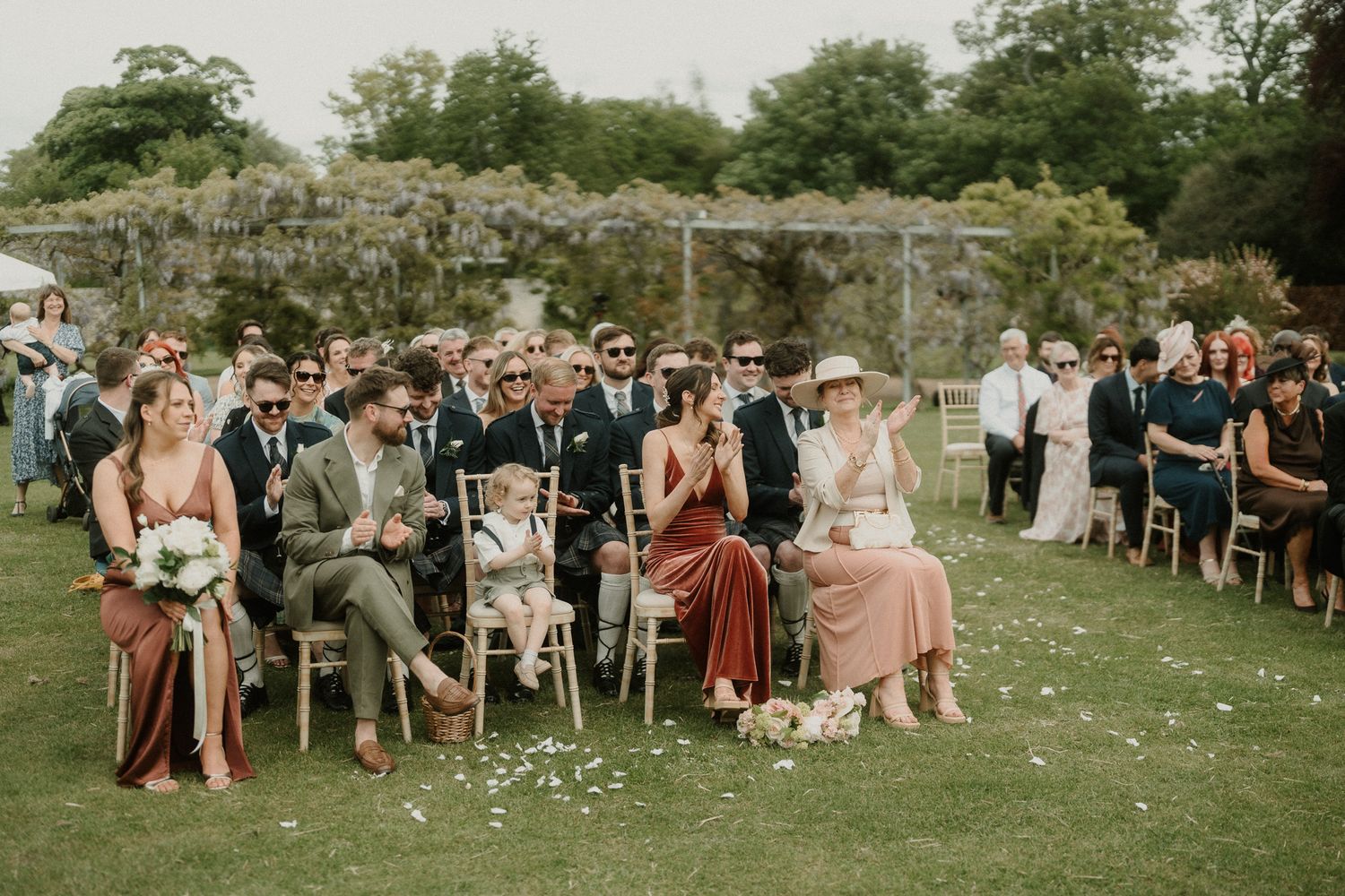 Guests seated outdoors on chairs at a garden wedding ceremony, surrounded by lush greenery and stone walls.