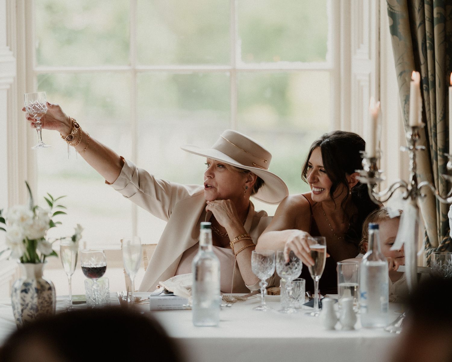 Two women laughing and raising glasses in a toast at an elegant wedding reception table with candles and glassware.