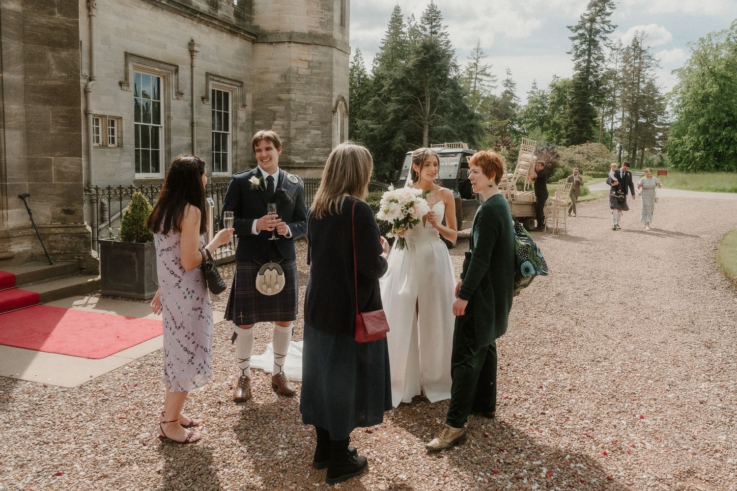Wedding guests gather outside a Scottish castle venue, bride holding bouquet, groom in tartan kilt, on a cloudy day.