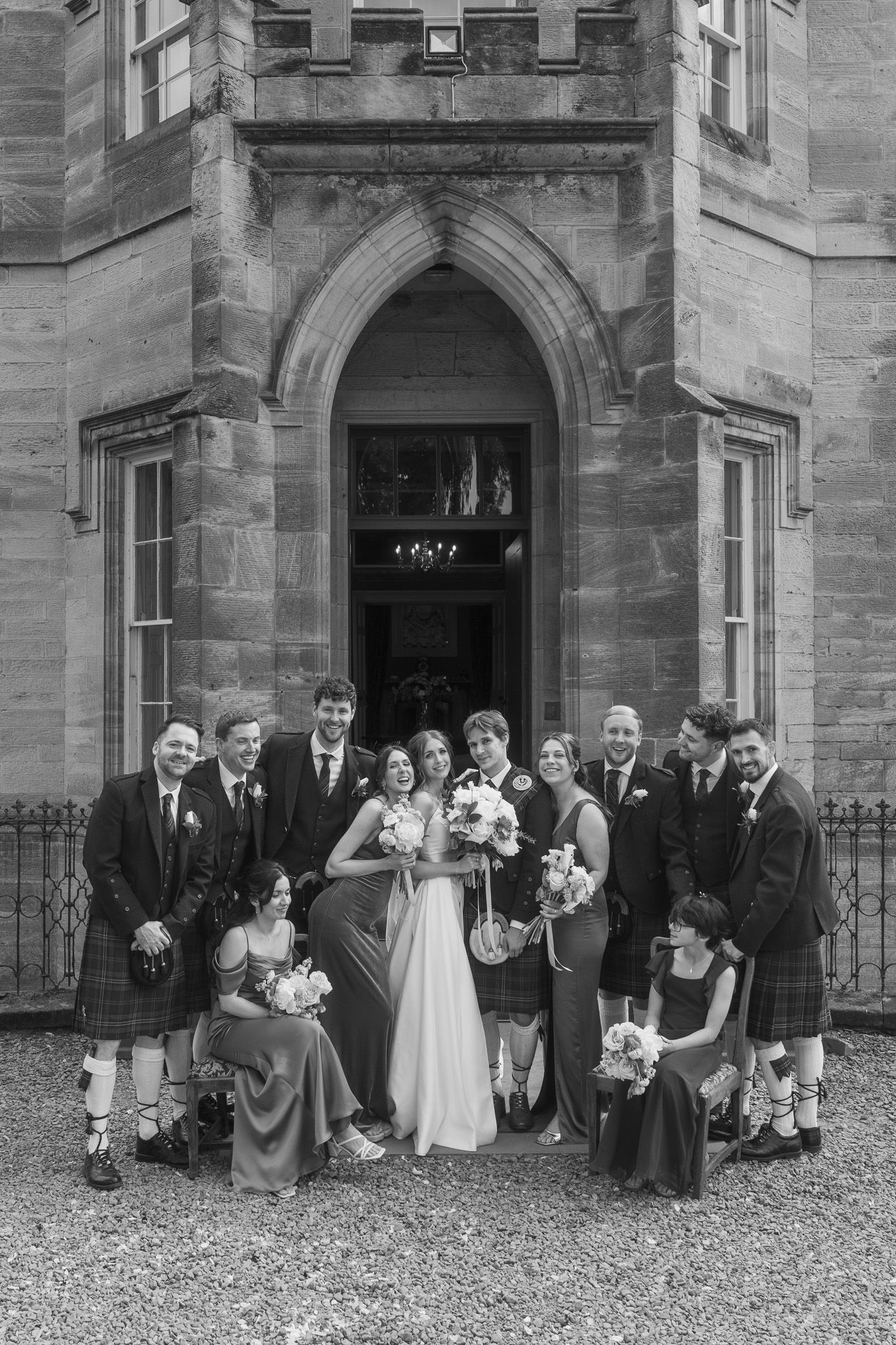 Black and white wedding party photo outside a historic stone building, bride and groom surrounded by guests in kilts and formal wear.