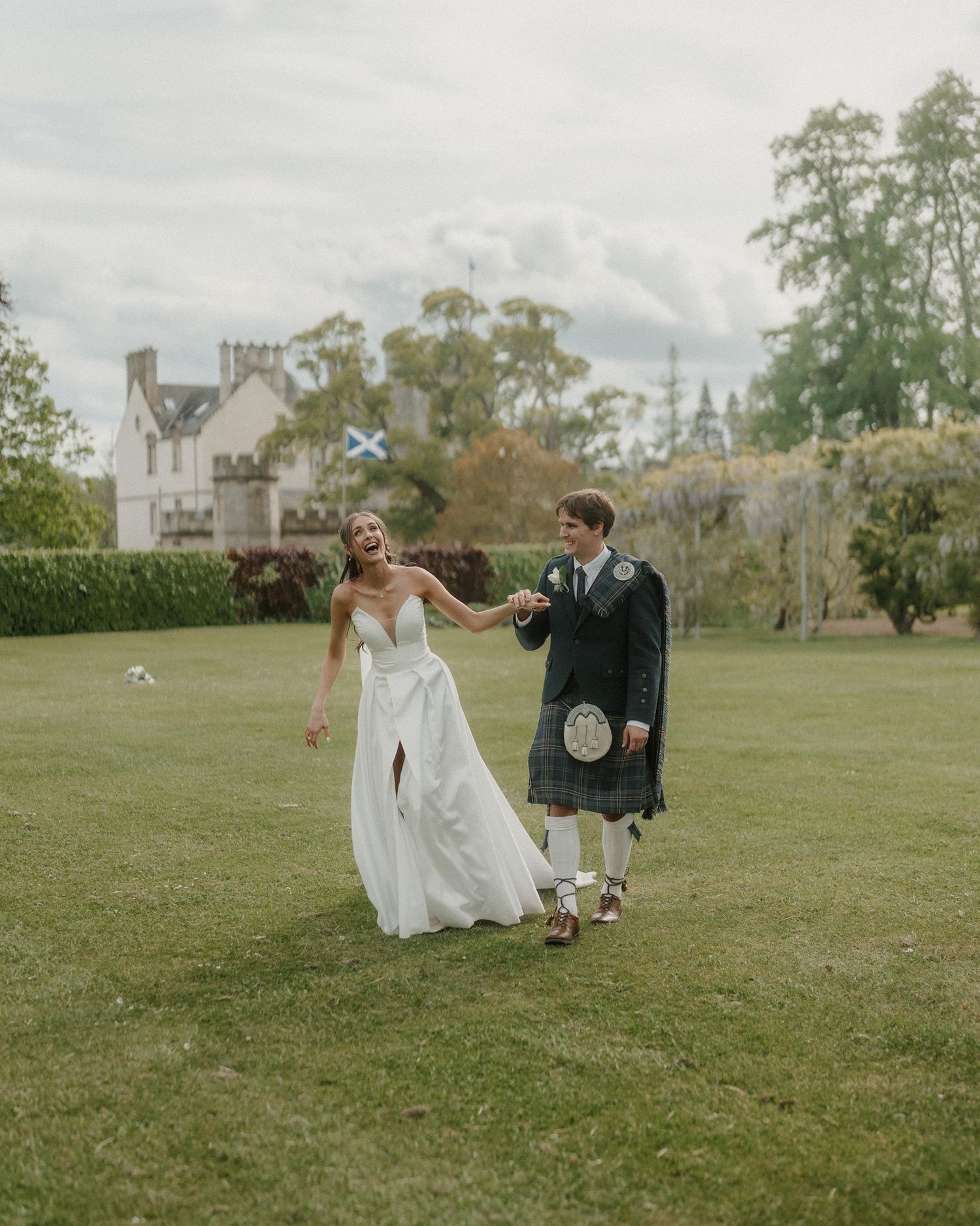 Bride and groom walking hand in hand on green lawn before Scottish castle, groom wearing traditional kilt.