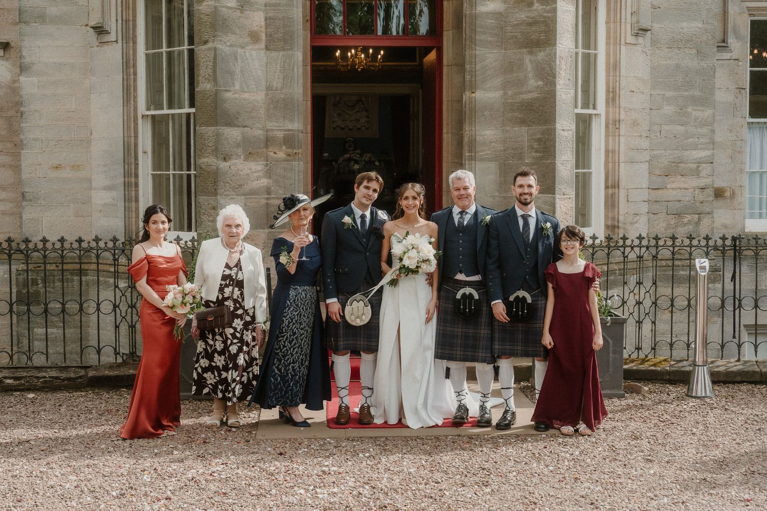 Wedding party posing outside a Scottish manor, groom in tartan kilt, bride in white gown, family in formal attire.