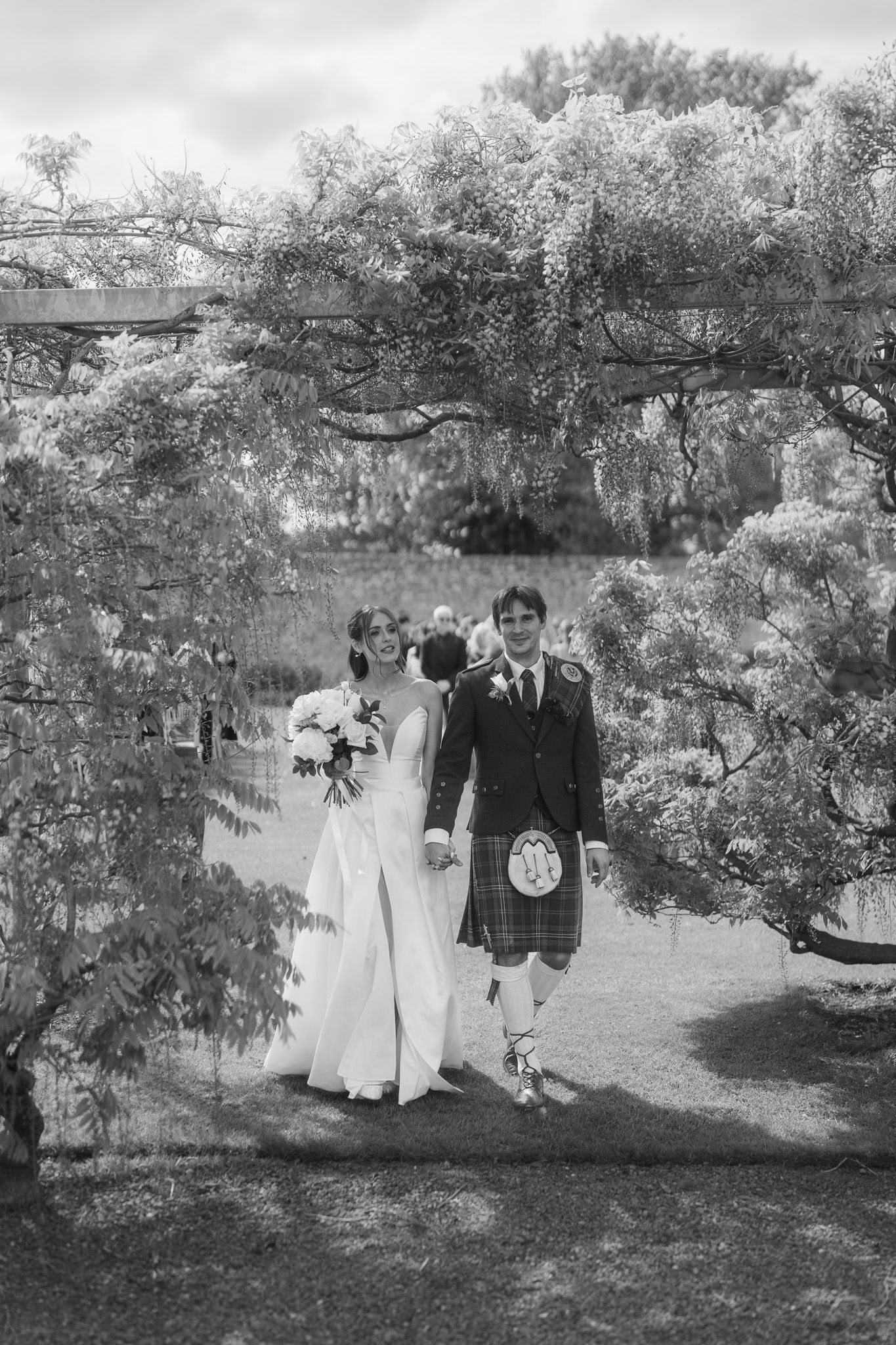 Black and white photo of a bride and groom in Scottish kilt walking hand in hand through a garden pergola.