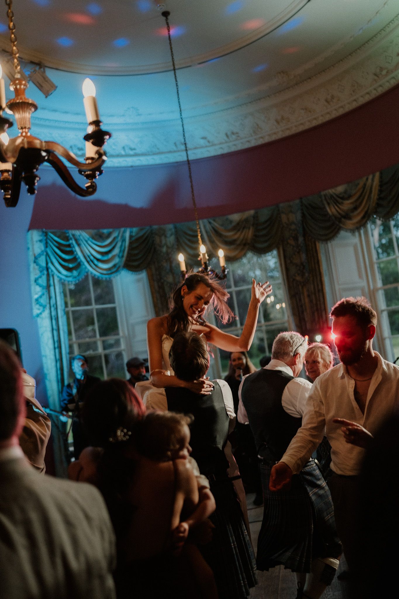 Guests dancing joyfully at an elegant wedding reception in a grand ballroom with chandelier and blue mood lighting.