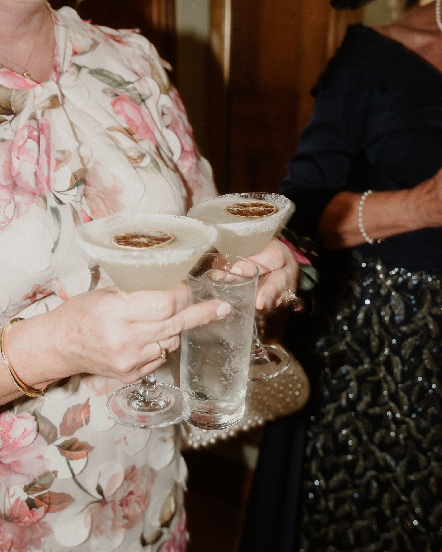 Woman in floral dress holding cocktails and water glass on tray at elegant party event.