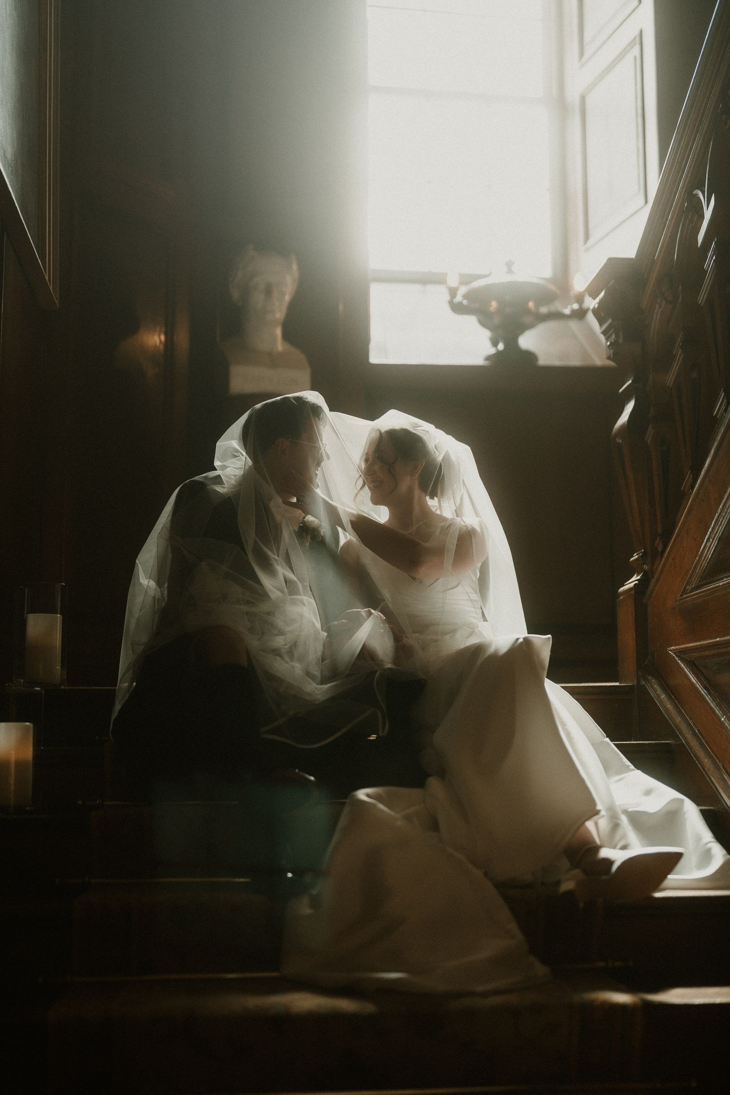 Bride and groom share an intimate moment under a veil on stairs, bathed in soft window light in a dark wood interior.