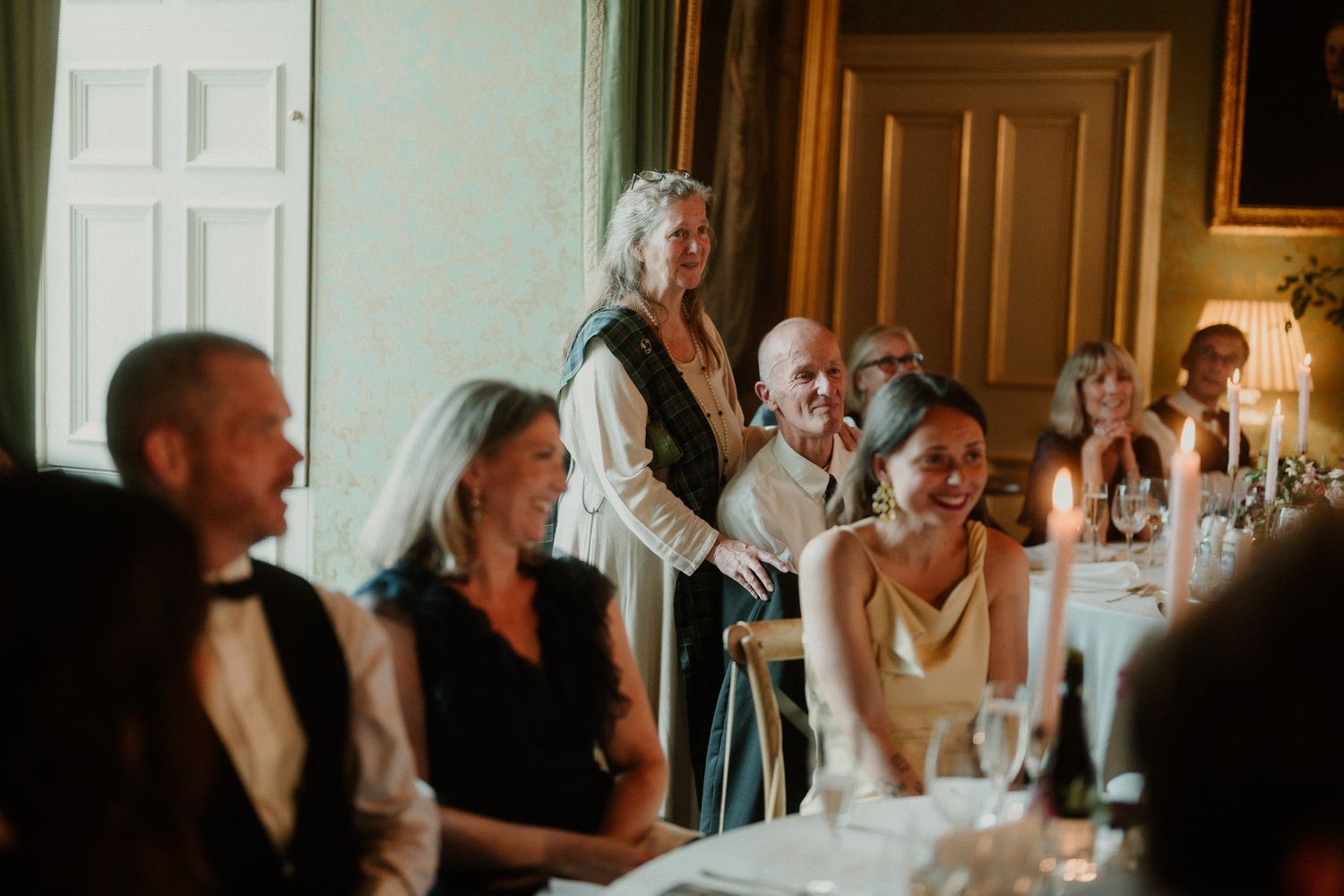 Guests in formal attire laugh and listen during a candlelit dinner reception as a woman stands to give a speech in an elegant room.