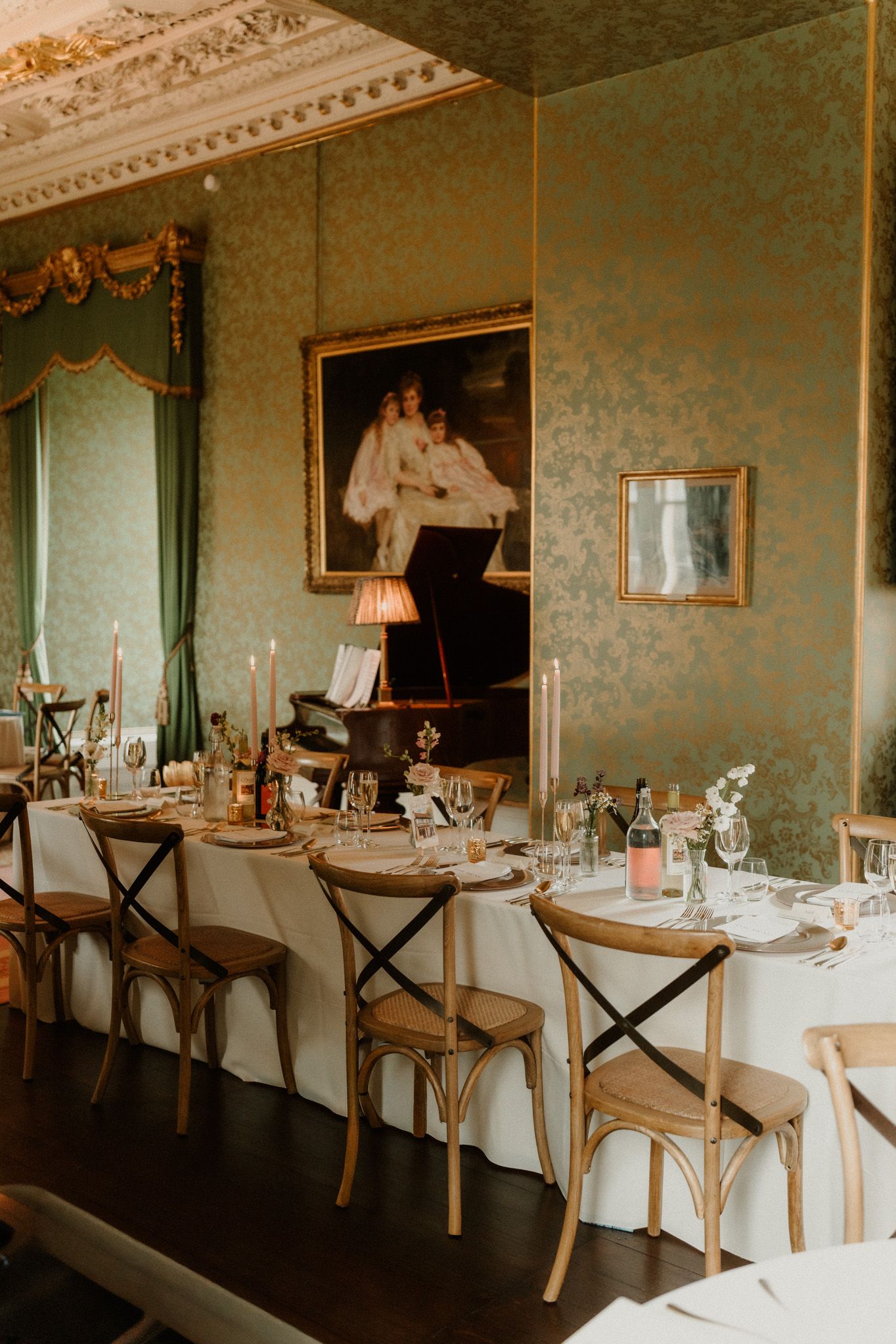 Elegant wedding reception room with a long table, cross-back chairs, candles, a grand piano and a framed portrait on green damask walls.