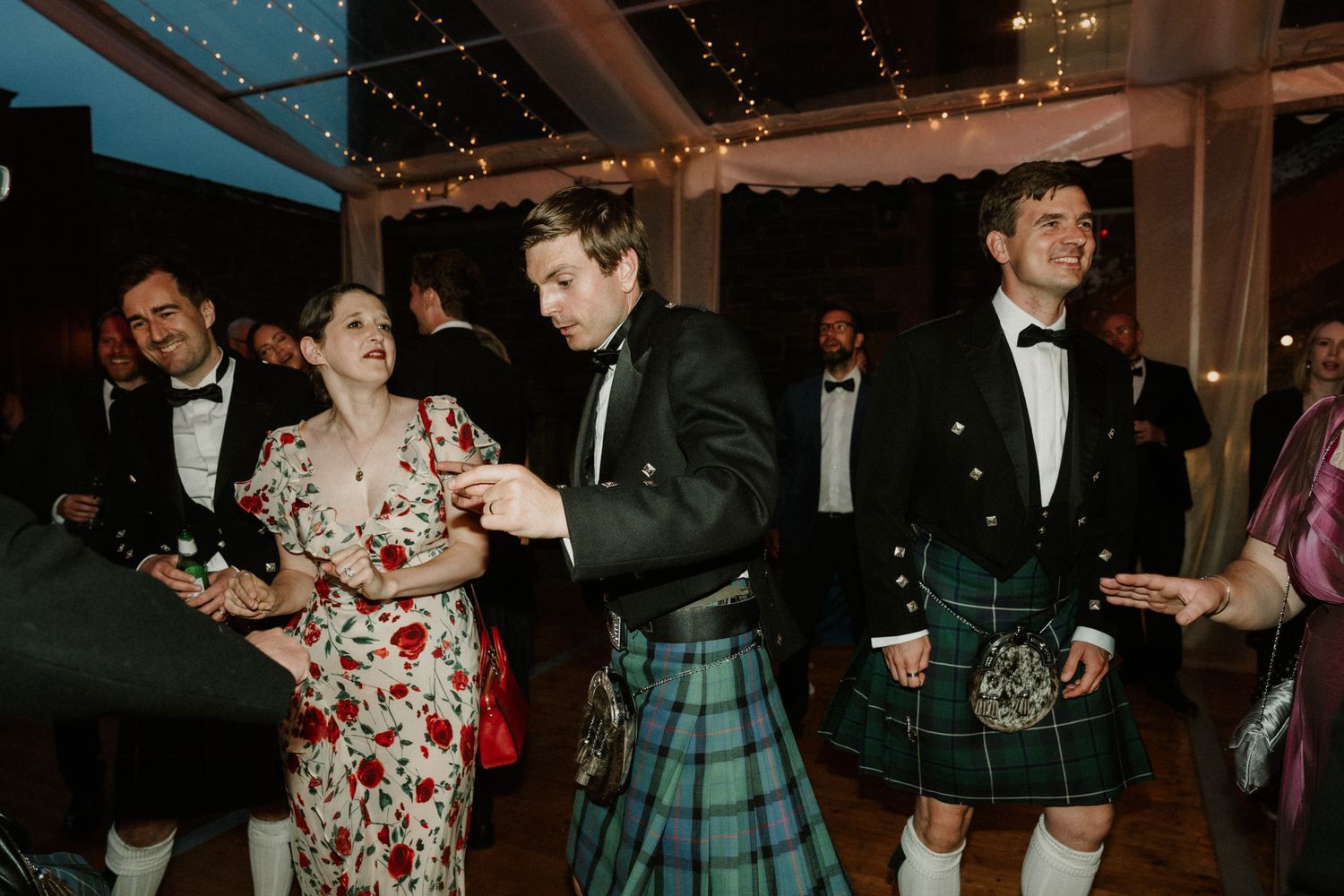 Guests in kilts and formal wear dance and laugh together at an evening wedding reception under a tent with string lights.
