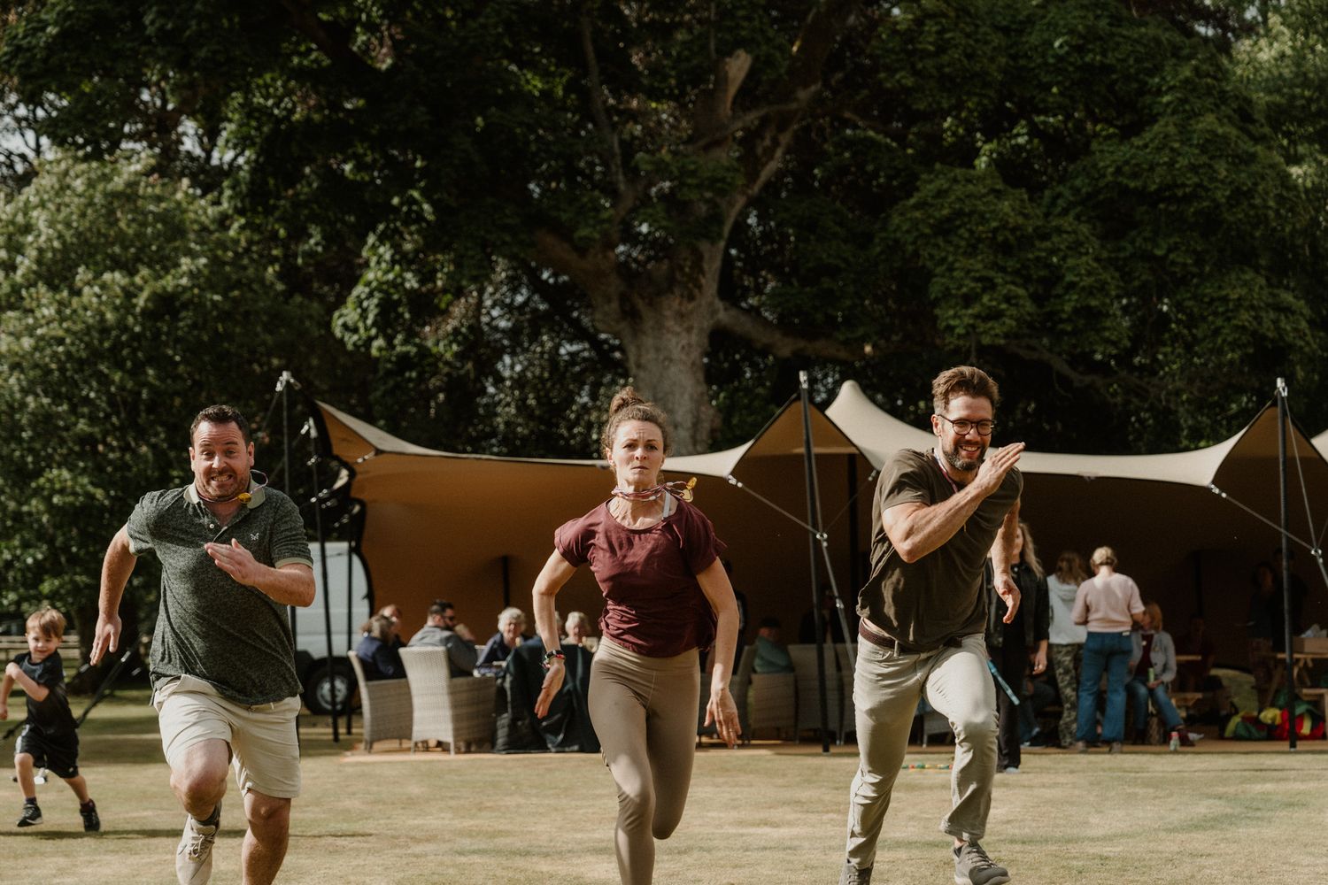 Three people run across a sunny grass field in casual clothes, with tents, tall trees, and onlookers in the background at an outdoor event.