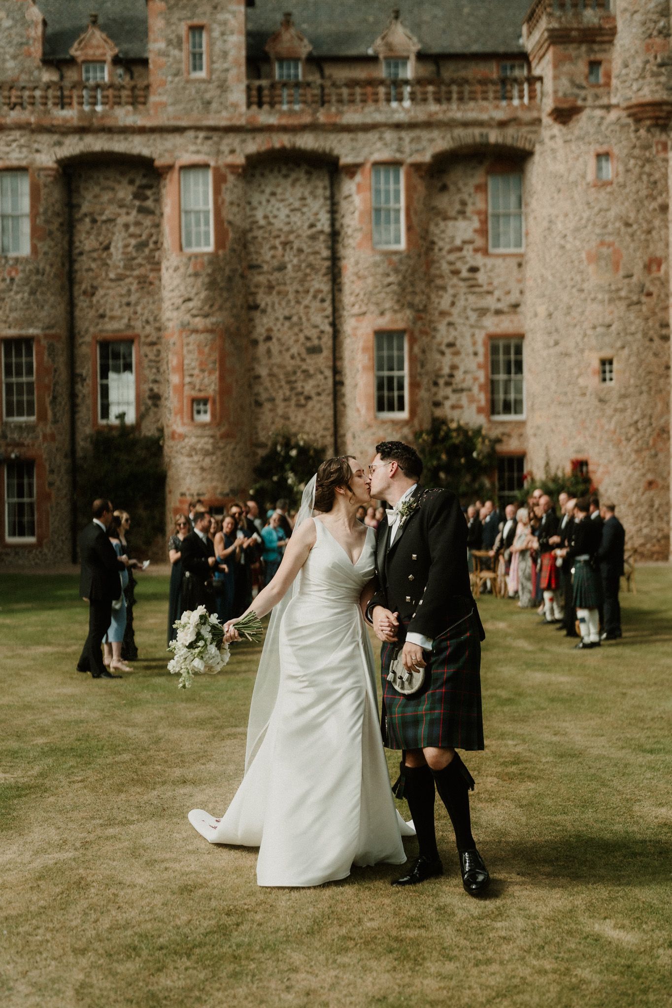Bride in a white gown and groom in a kilt share a kiss on the lawn before a stone castle as wedding guests look on.