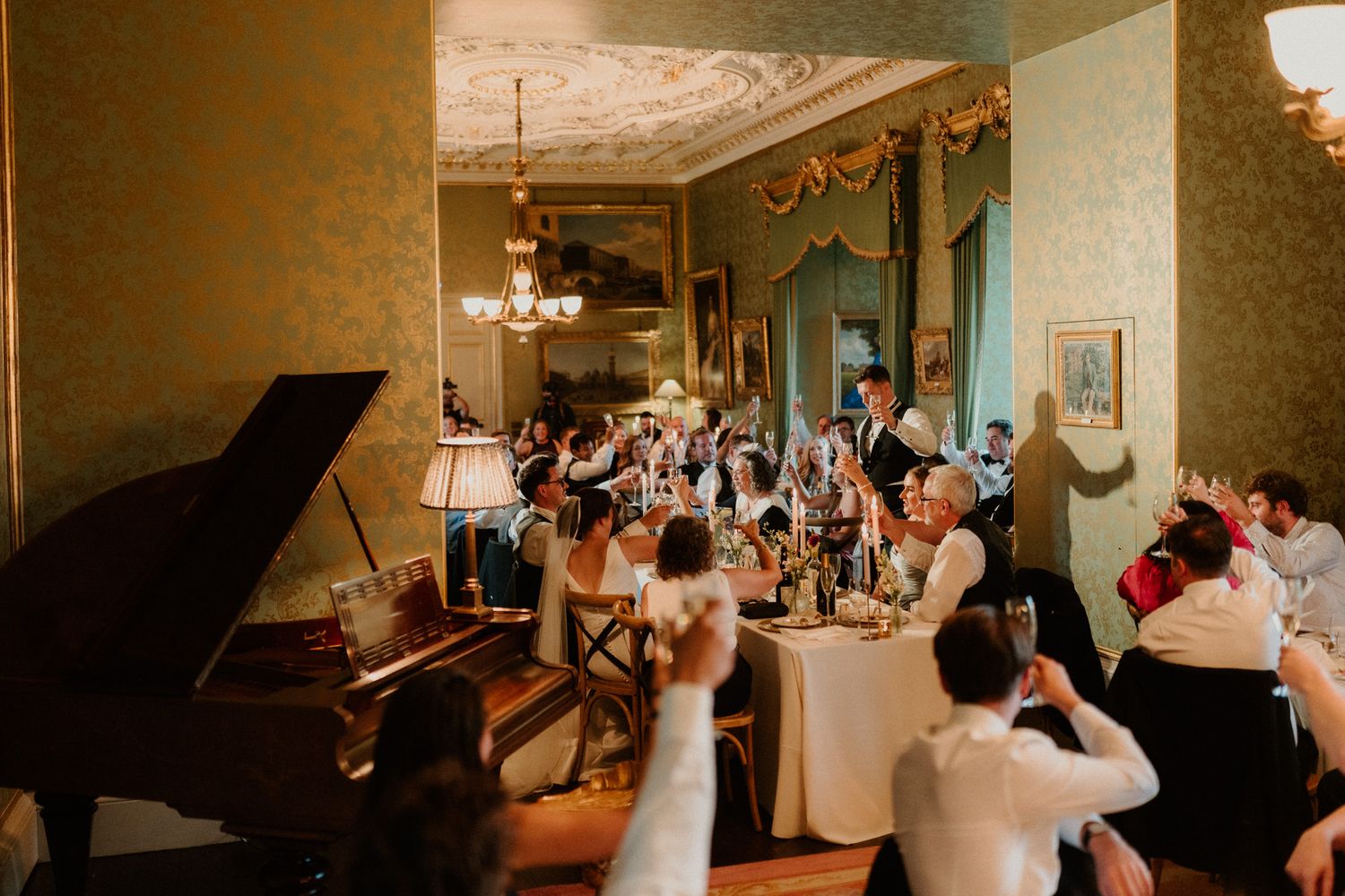 Wedding guests raise glasses in a toast around a candlelit table in an elegant ballroom with a grand piano and ornate decor.