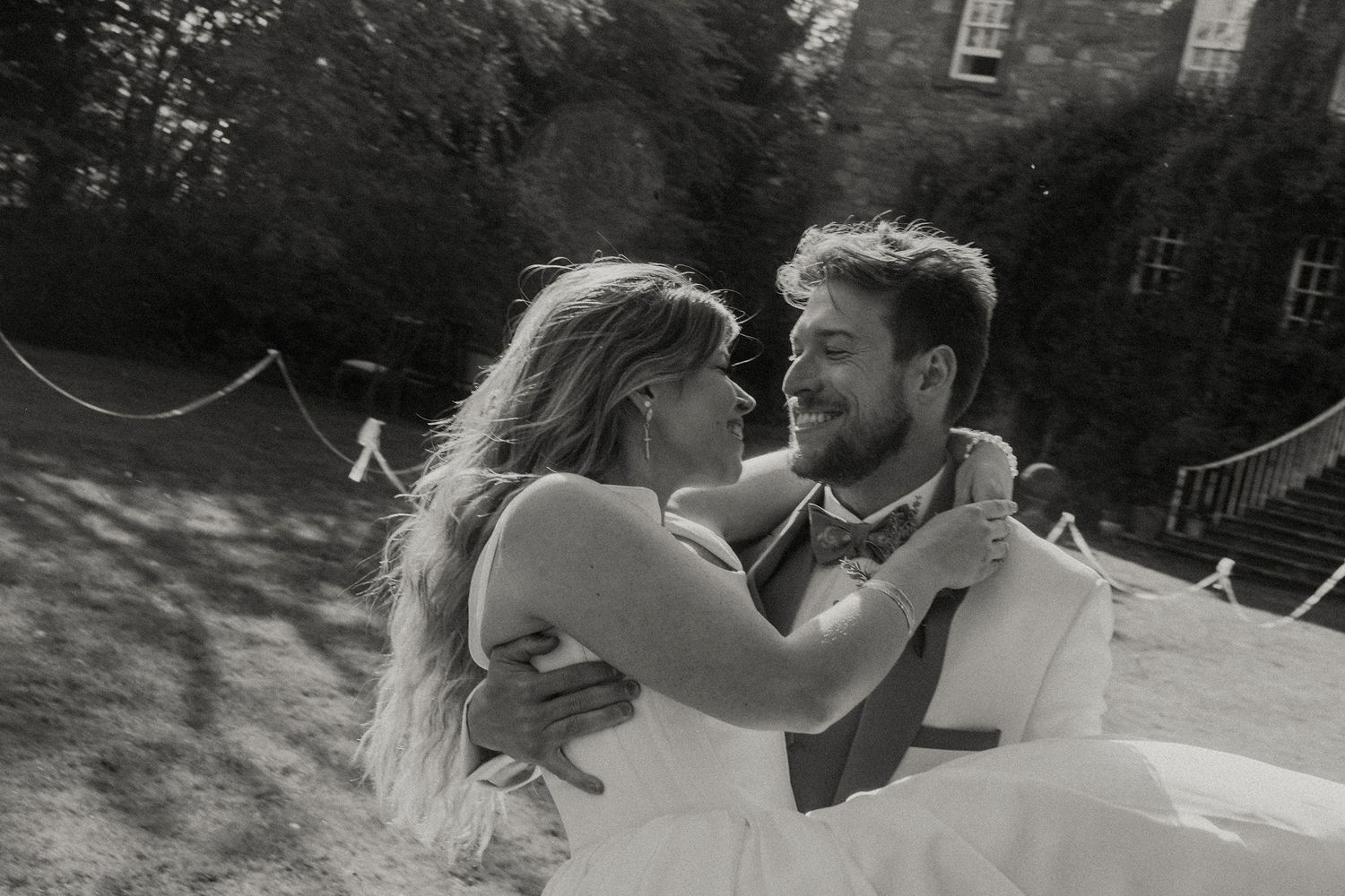 Black and white photo of a smiling bride and groom embracing outdoors at a wedding venue surrounded by trees.