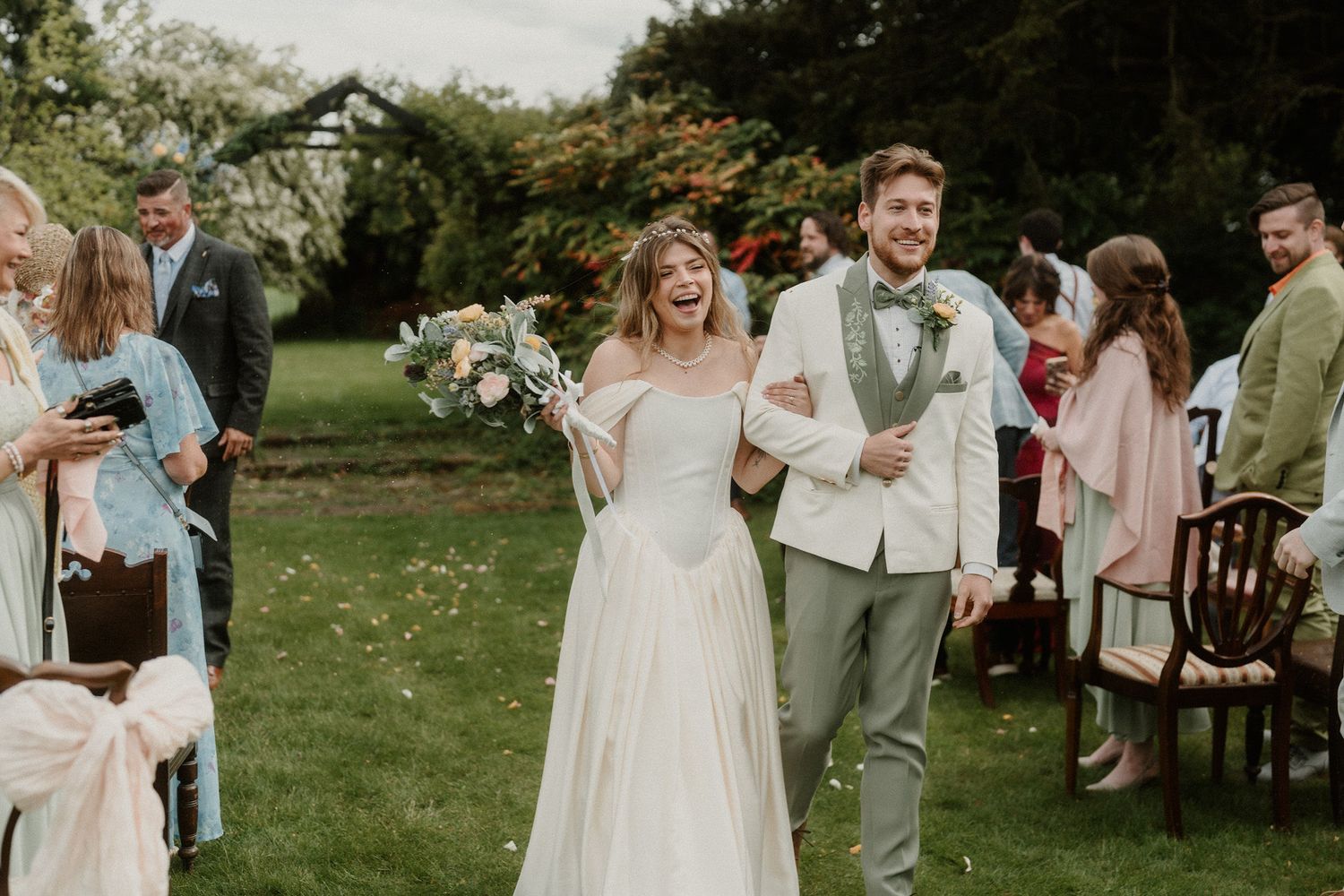Happy newlywed couple walking down outdoor wedding aisle, bride in white gown holding bouquet, groom in grey suit.