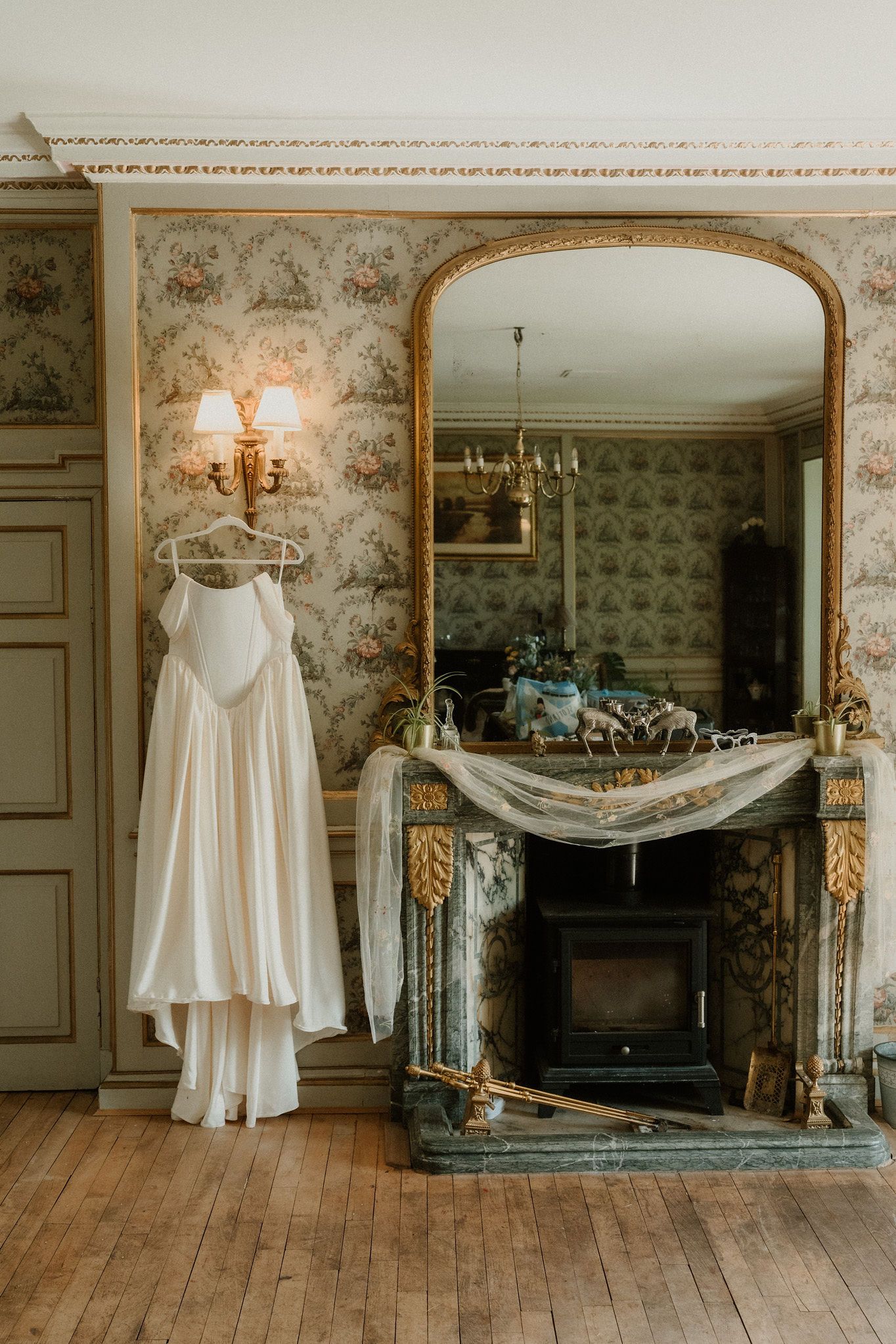 Elegant white wedding dress hanging near a ornate gold fireplace mirror in a vintage French-style room with floral wallpaper.