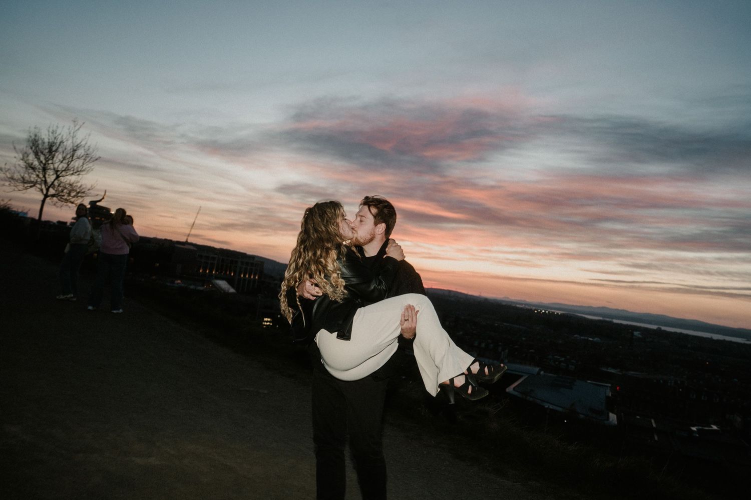 Couple sharing a romantic kiss at sunset, man carrying woman in his arms against a colorful twilight sky.