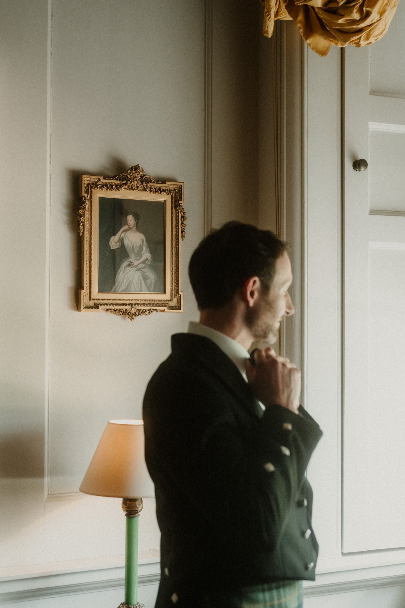 Groom in Scottish attire adjusting his bow tie beside a vintage portrait and lamp in an elegant room.