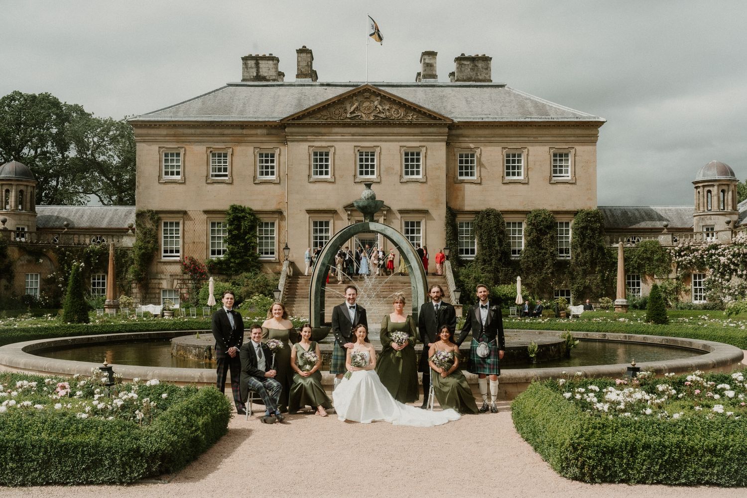 Wedding party posing in formal garden with ornate Georgian manor house backdrop, bride in white gown surrounded by guests.