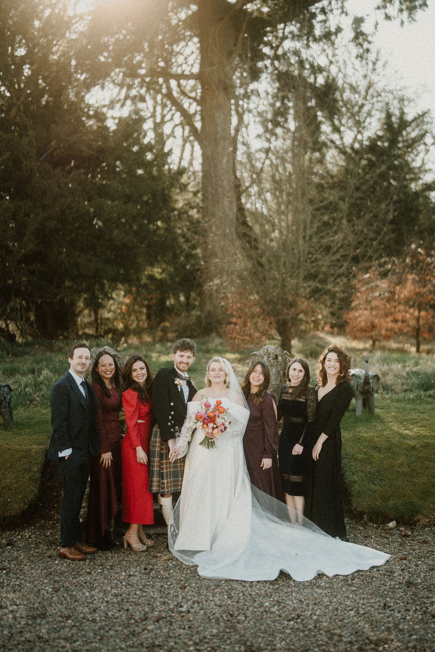 Wedding party in formal attire poses together on gravel path during autumn outdoor celebration.