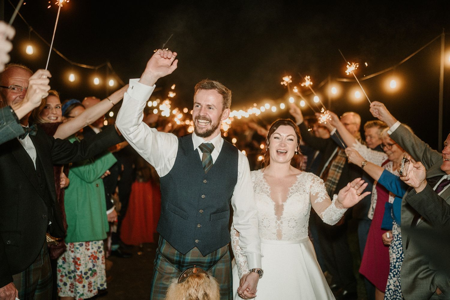 Wedding guests hold sparklers creating a magical nighttime celebration atmosphere under string lights.
