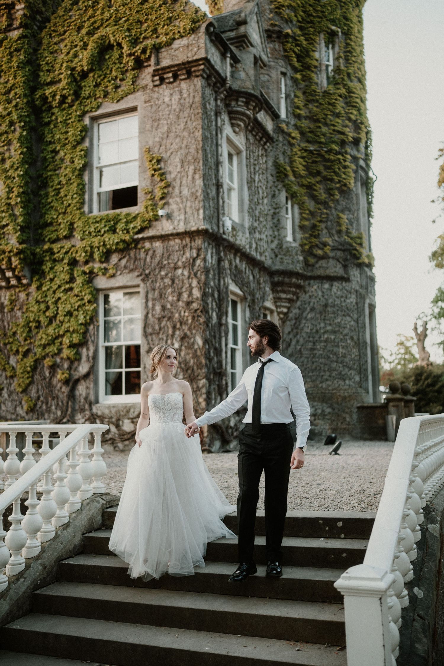 A couple poses on white stone steps leading to an ivy-covered historic building.