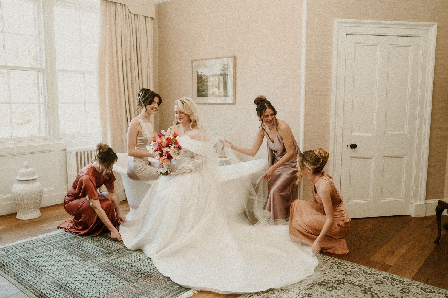 Wedding party members assist with the bride's dress preparations in an elegant room.
