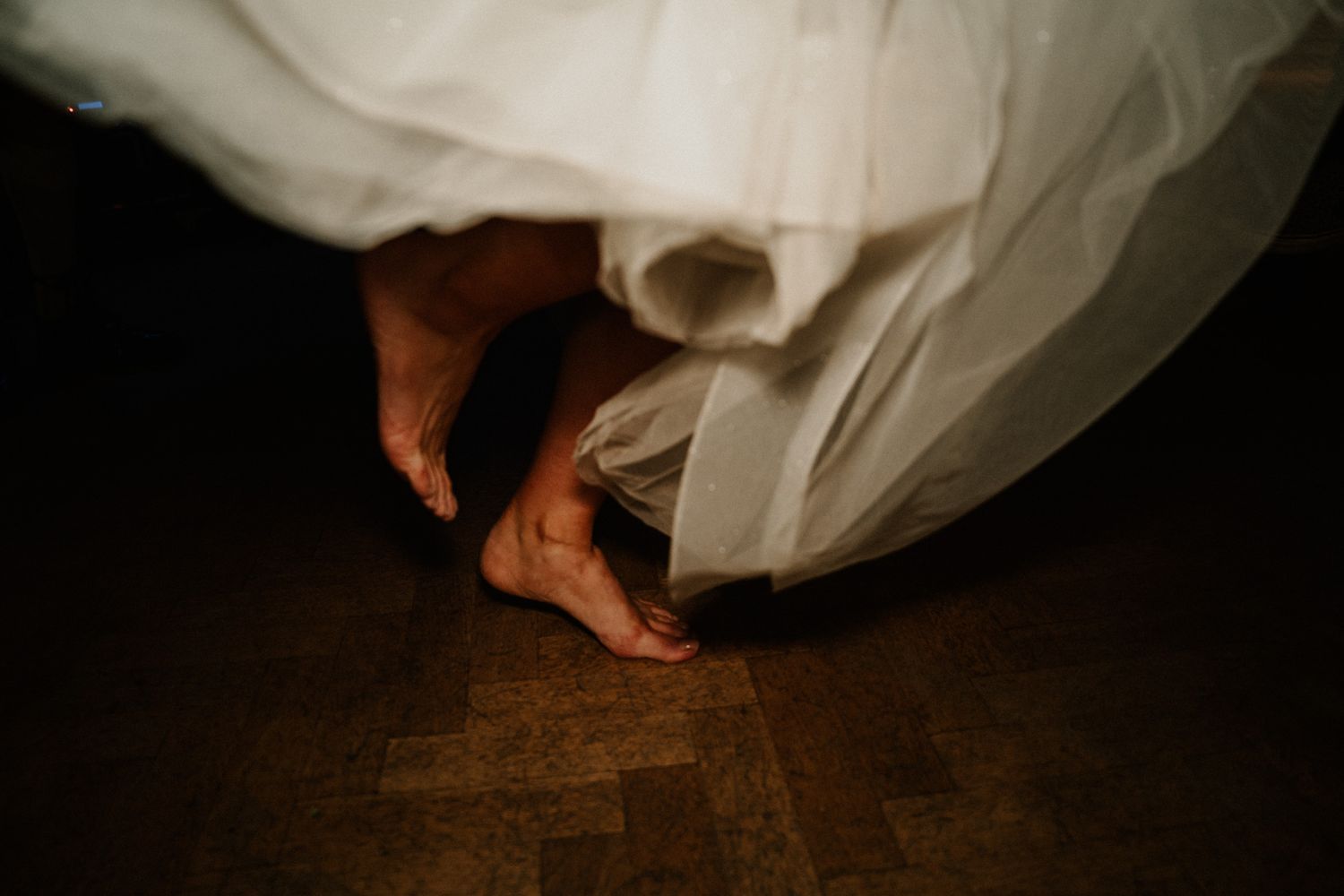 Bare feet peek out from under a flowing white wedding dress against a dark wooden floor.