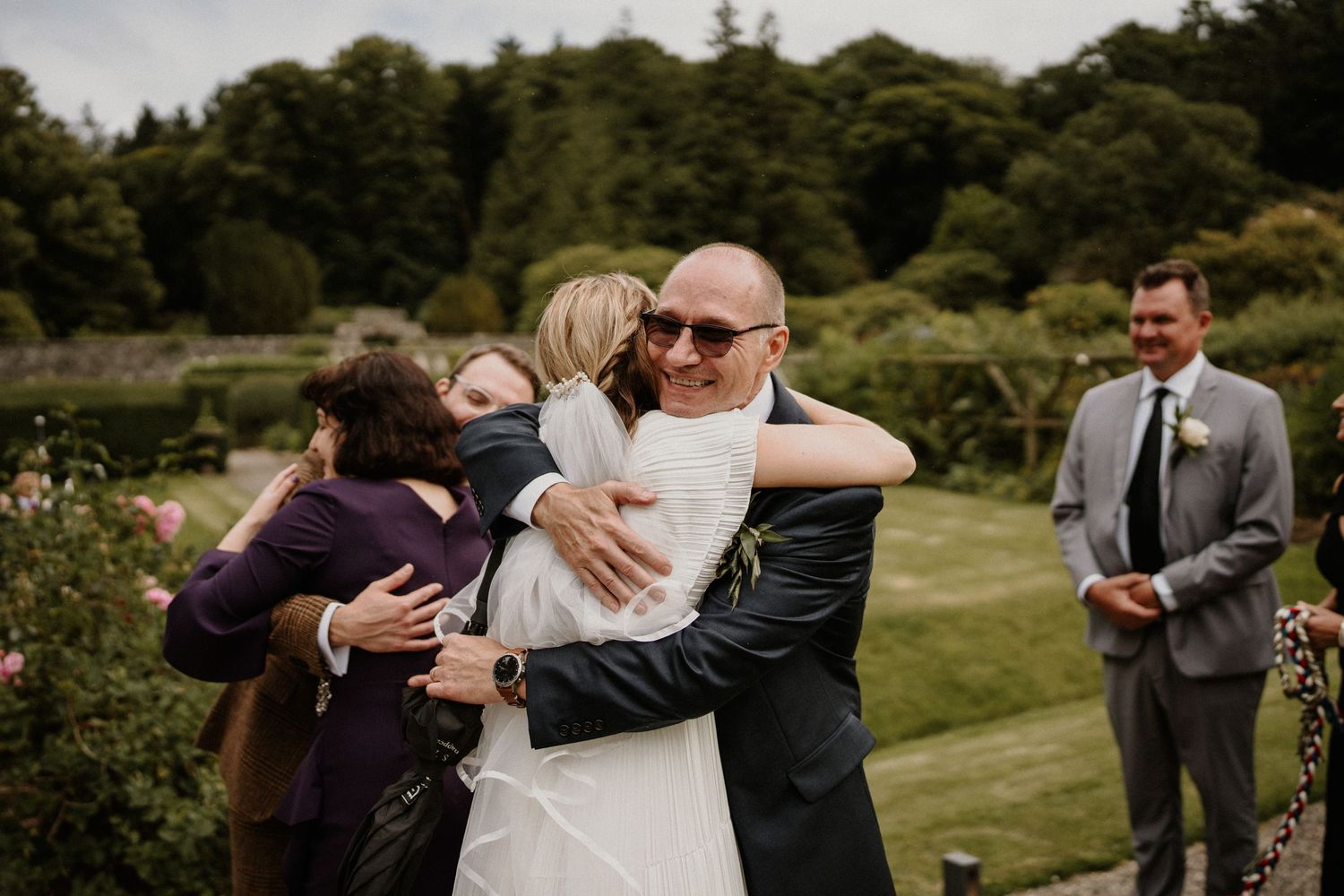 Wedding guests share emotional embraces on a manicured garden lawn during the celebration.