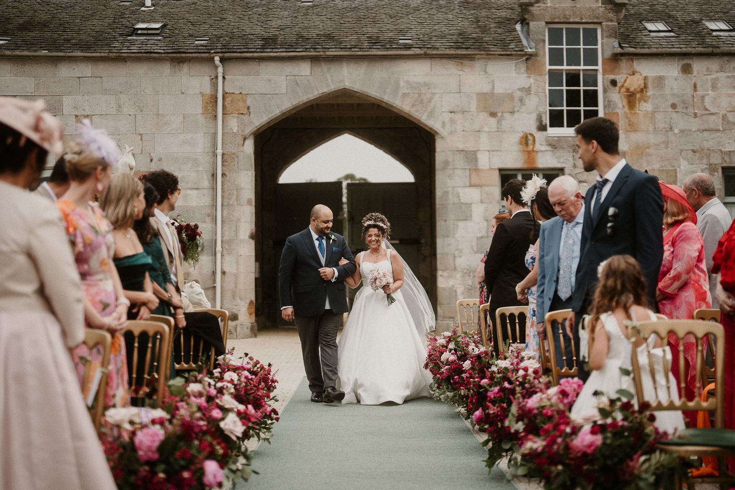 Stone castle courtyard wedding ceremony with burgundy floral aisle decorations.