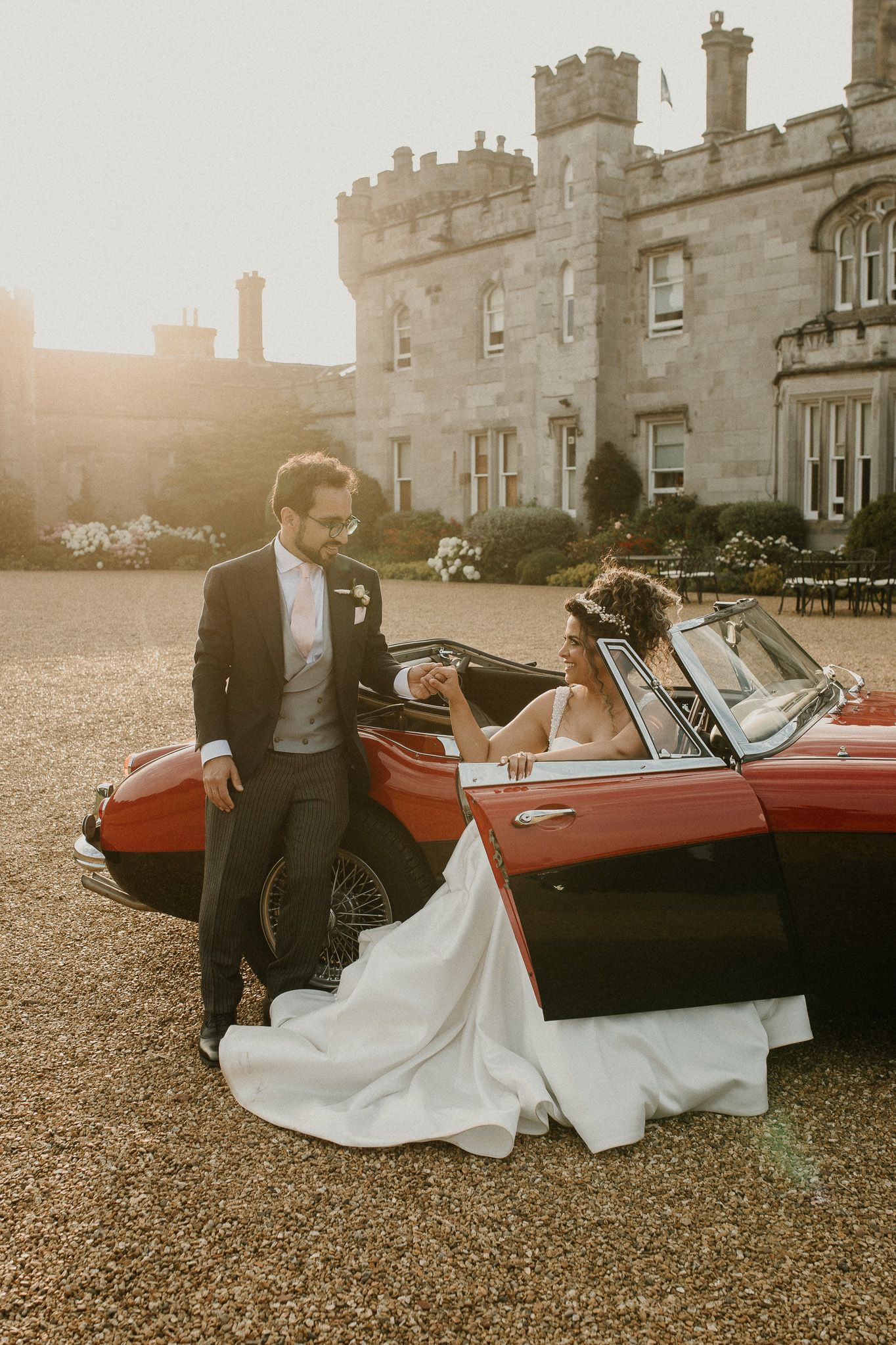 Wedding couple posing with a classic red sports car outside a majestic stone castle.