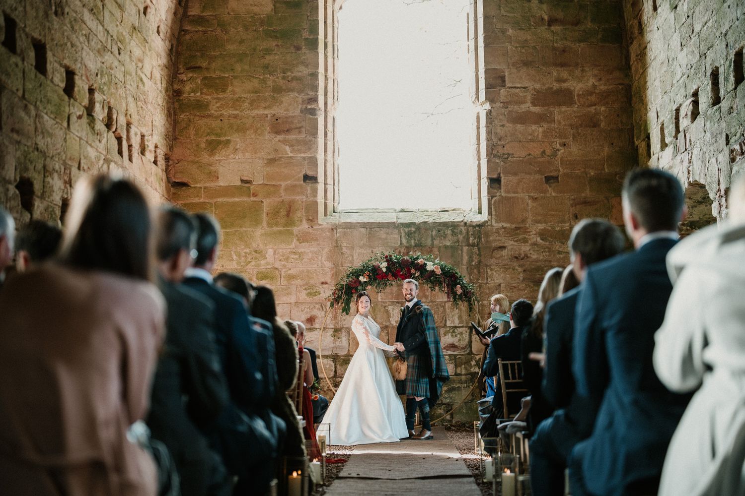 Romantic wedding ceremony under a floral arch in a historic stone chapel filled with guests.